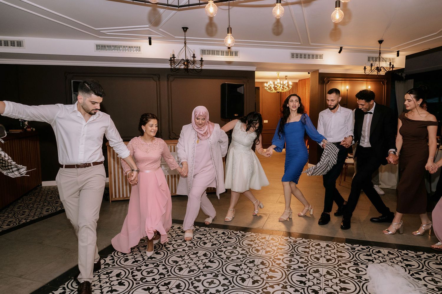 Wedding guests dance in a line formation on a patterned floor during the reception celebration.