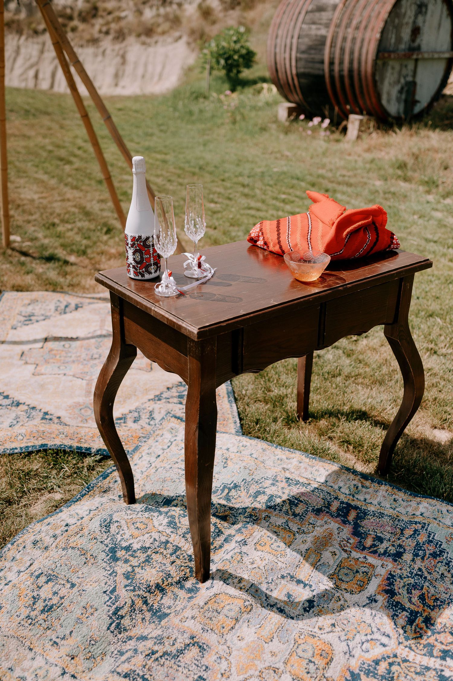 Antique side table with wine bottle and coral cloth set on patterned rug outdoors.