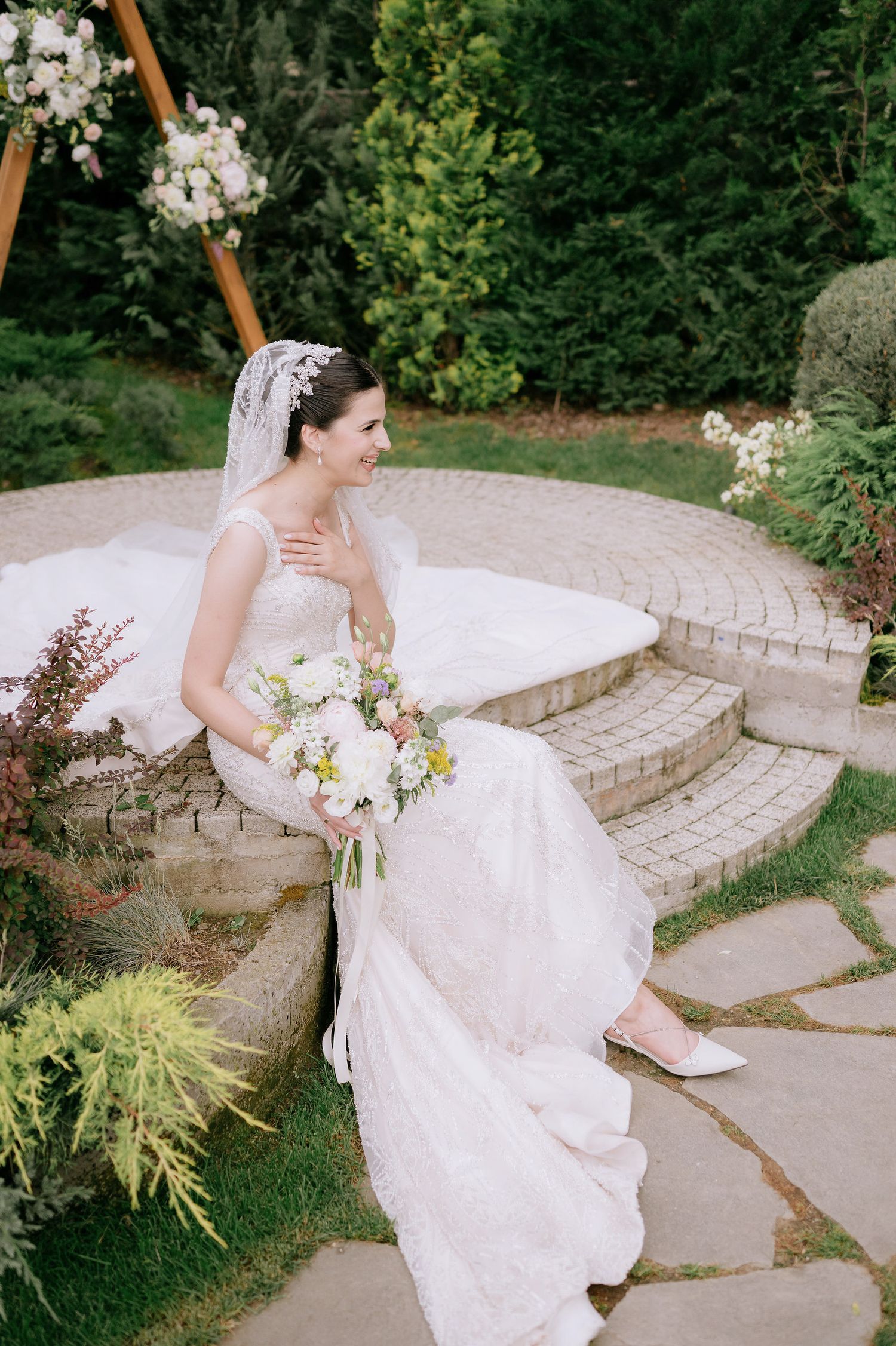 Bride in flowing white gown seated gracefully on curved stone patio steps.