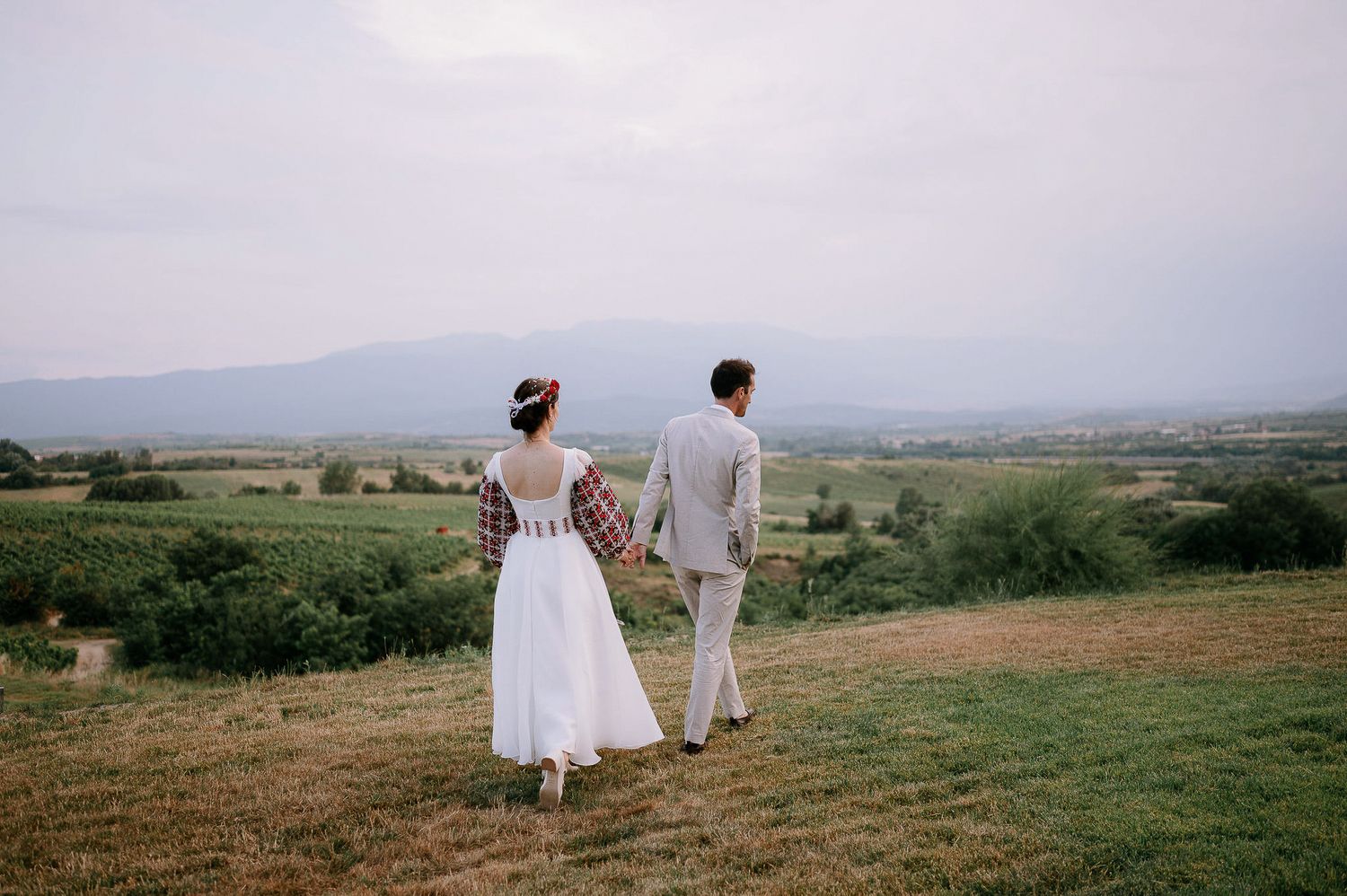 Newlyweds walk together on hilltop overlooking scenic mountain valley.