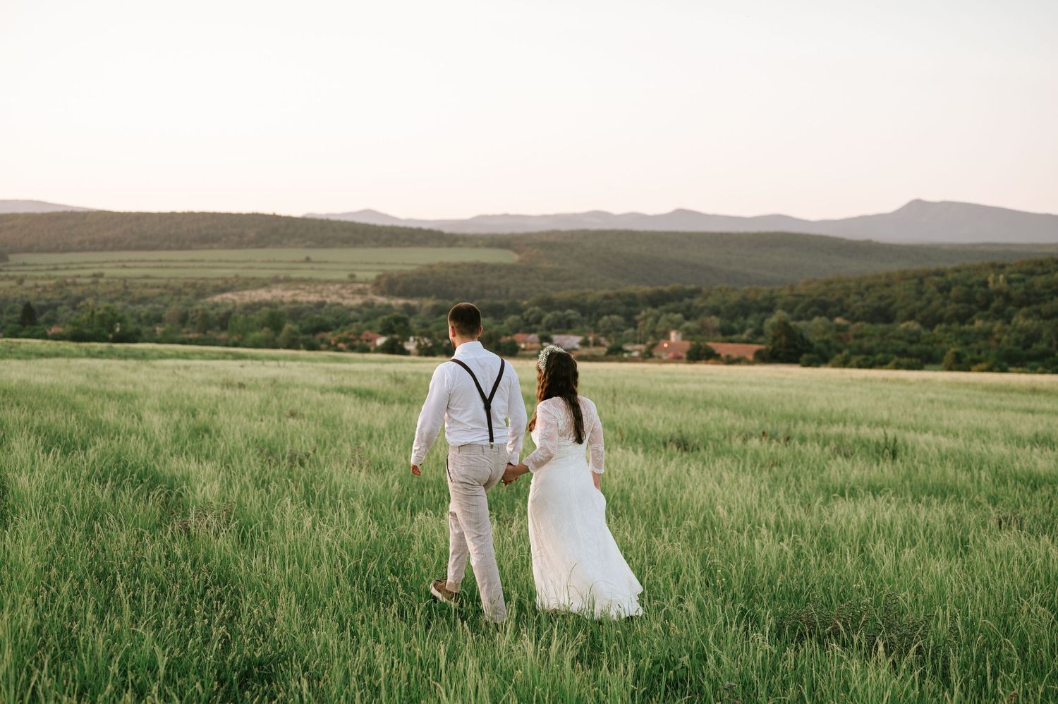 A wedding couple walks hand in hand through a scenic mountain meadow at sunset.