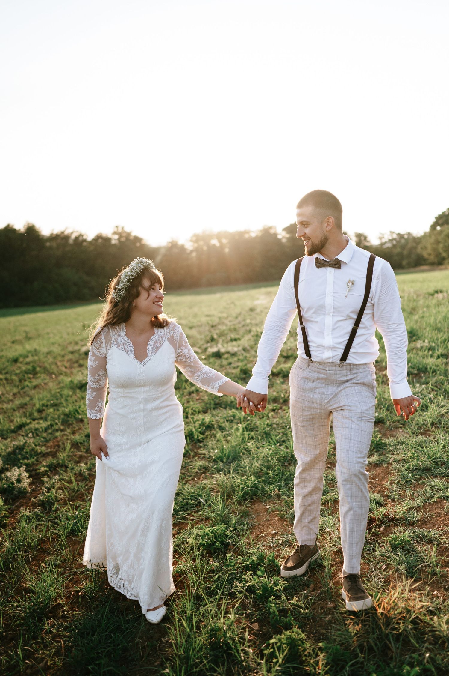 Newlyweds walking hand in hand through a sunlit meadow at golden hour in rustic wedding attire.