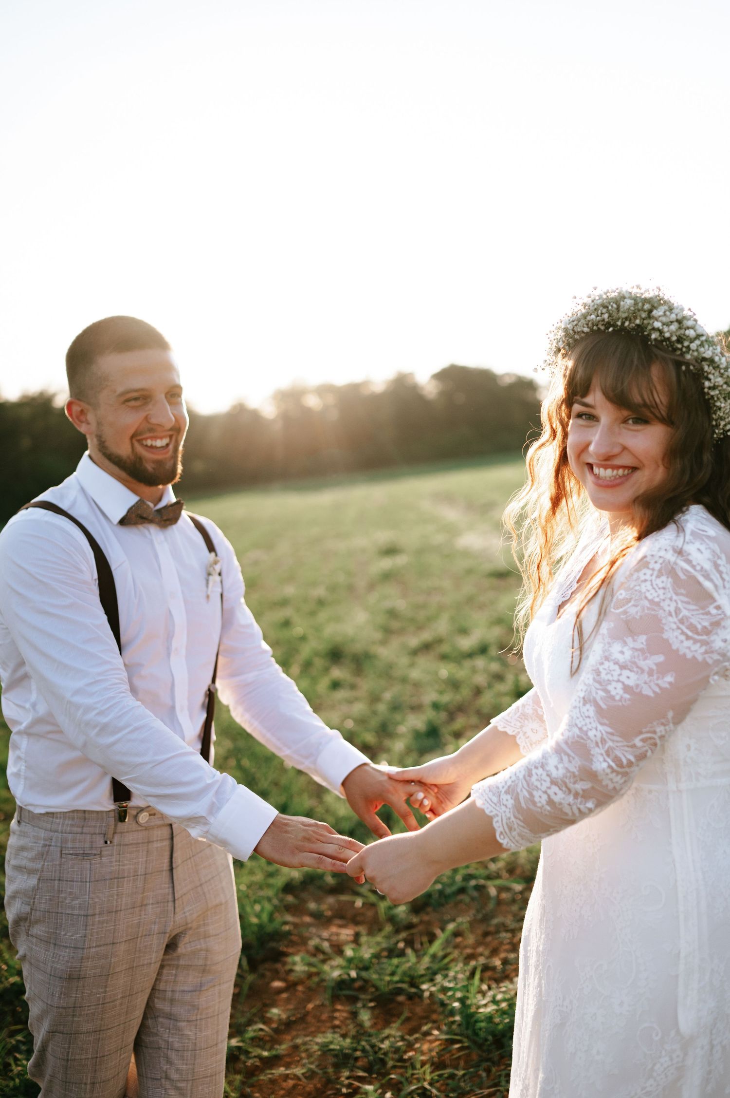 A romantic wedding couple holds hands in a sunlit field, with the bride in lace and groom in suspenders.