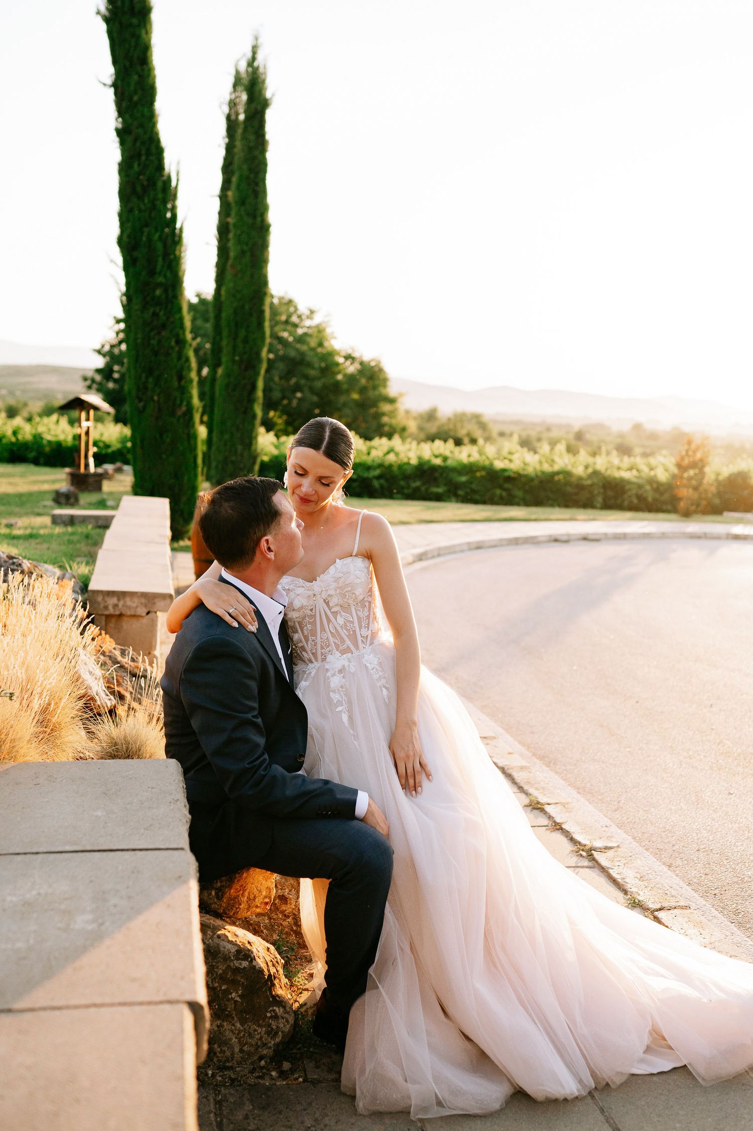 Elegant bride in lace gown and groom sharing intimate moments by cypress trees.