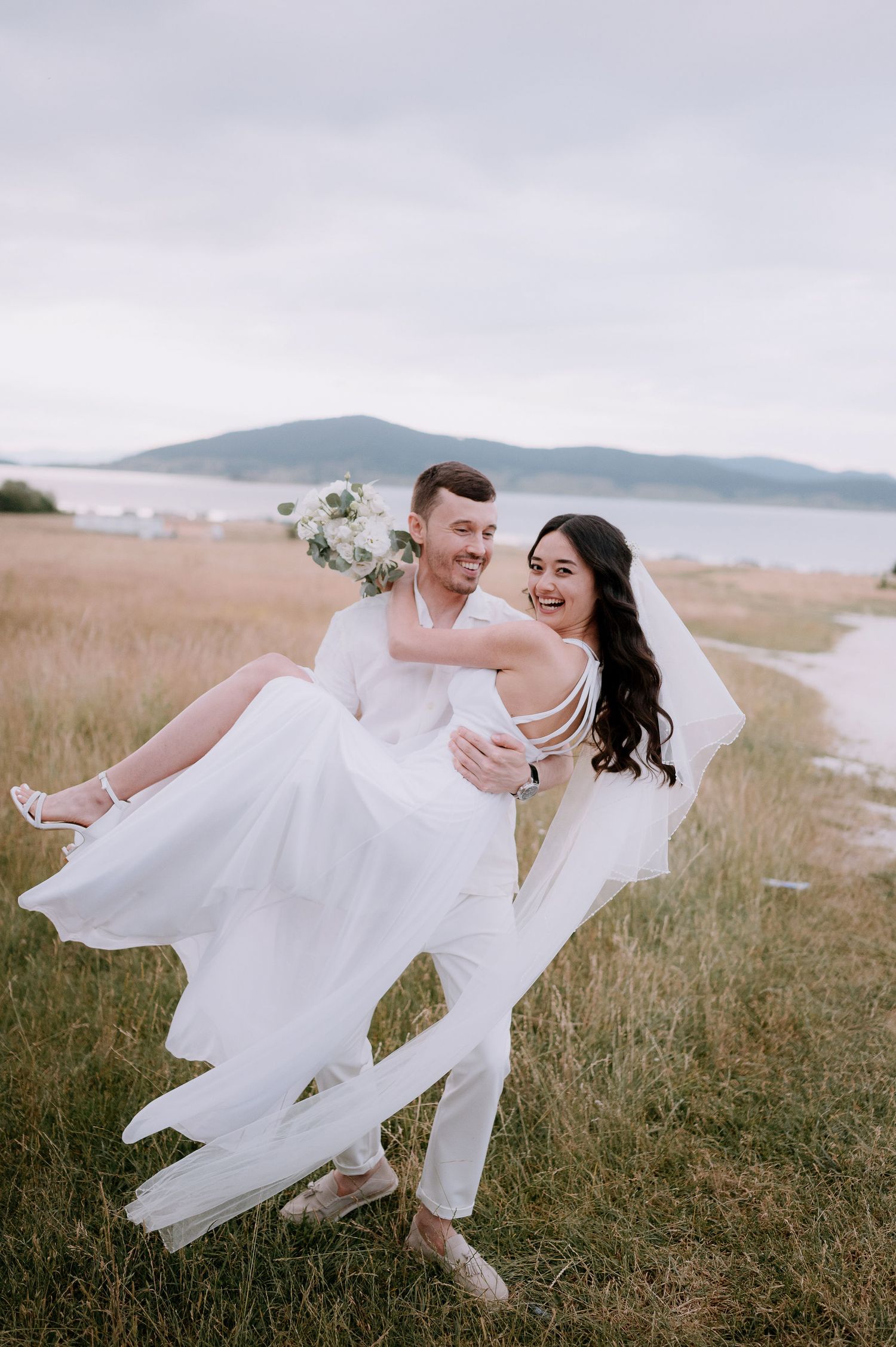 Groom carries bride in flowing white dress against coastal landscape backdrop.