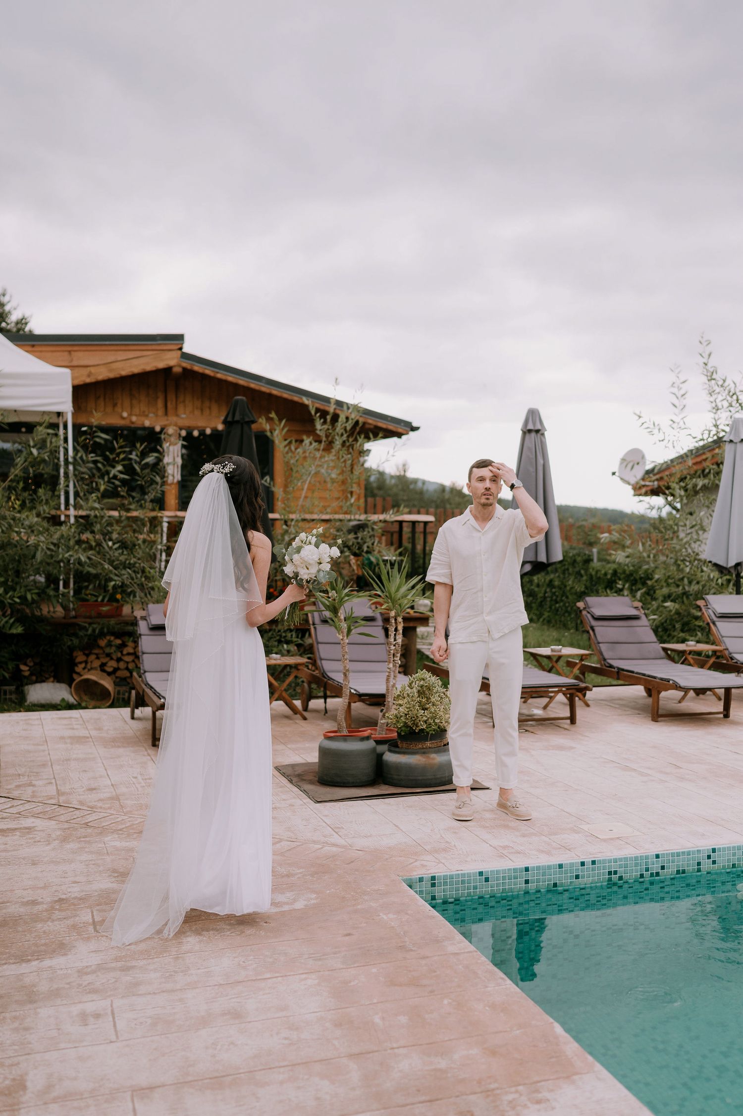 A wedding celebration takes place on a stone patio next to a turquoise pool with lounge chairs.
