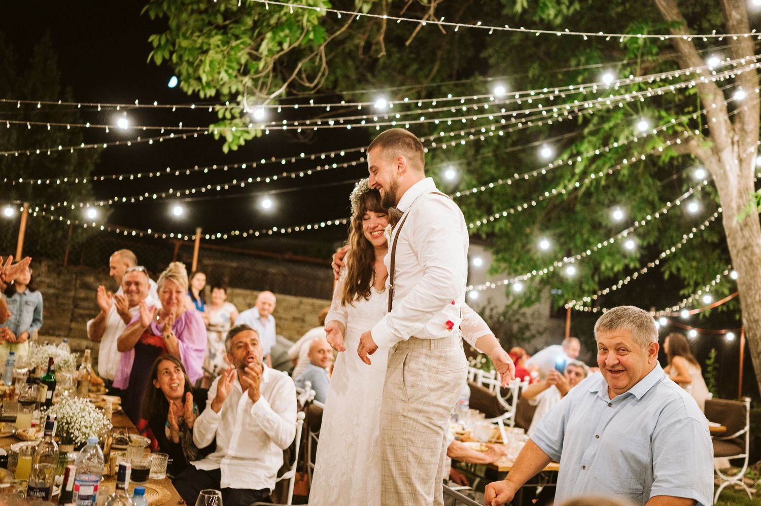 An outdoor wedding reception under twinkling string lights with guests celebrating at tables.