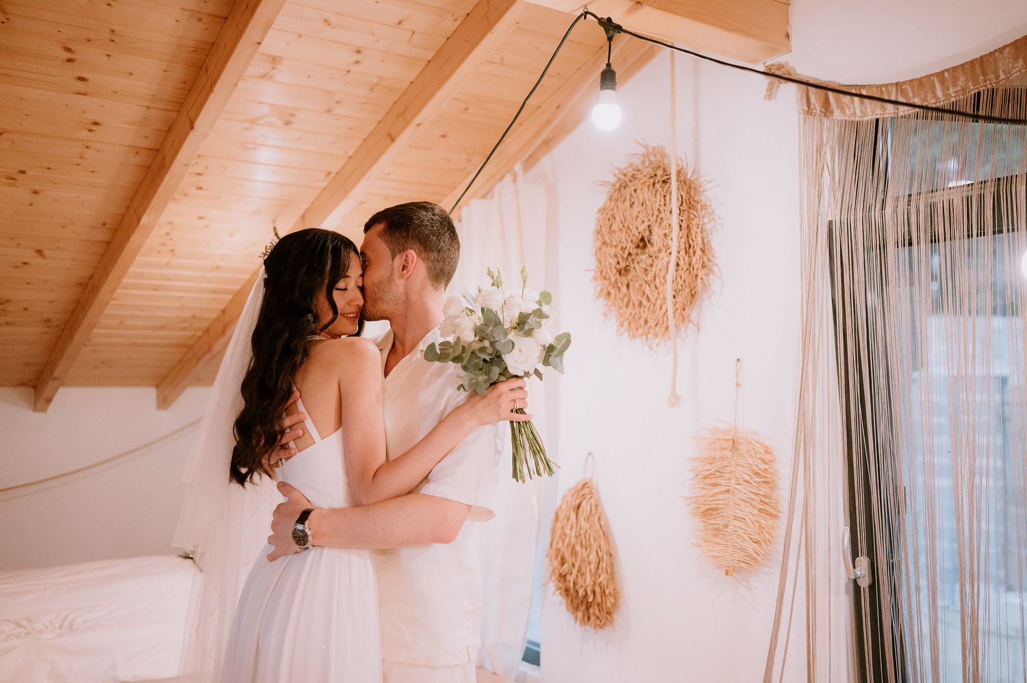 A couple dancing in a sunlit room with boho decor and wooden ceiling beams.