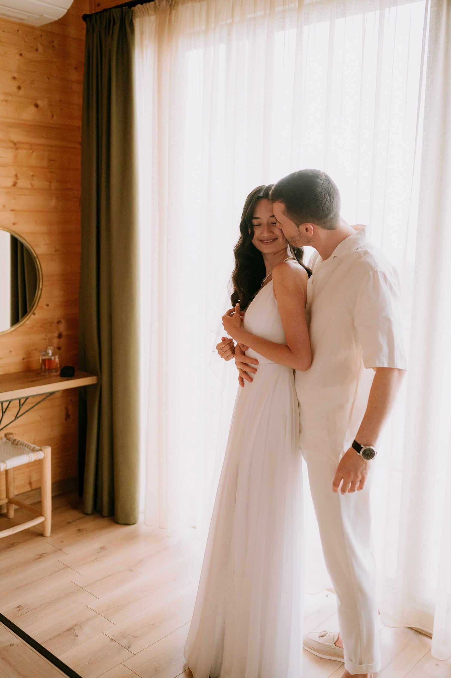 A romantic moment between a couple in white attire by a sunlit window in a wooden cabin.