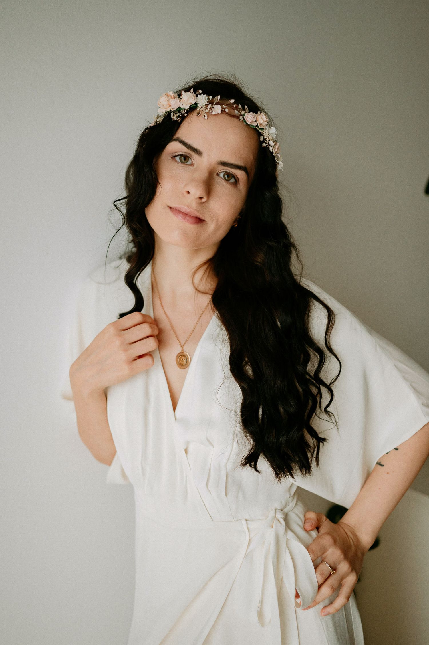 Ethereal bridal portrait in white robe with delicate floral crown against neutral background.