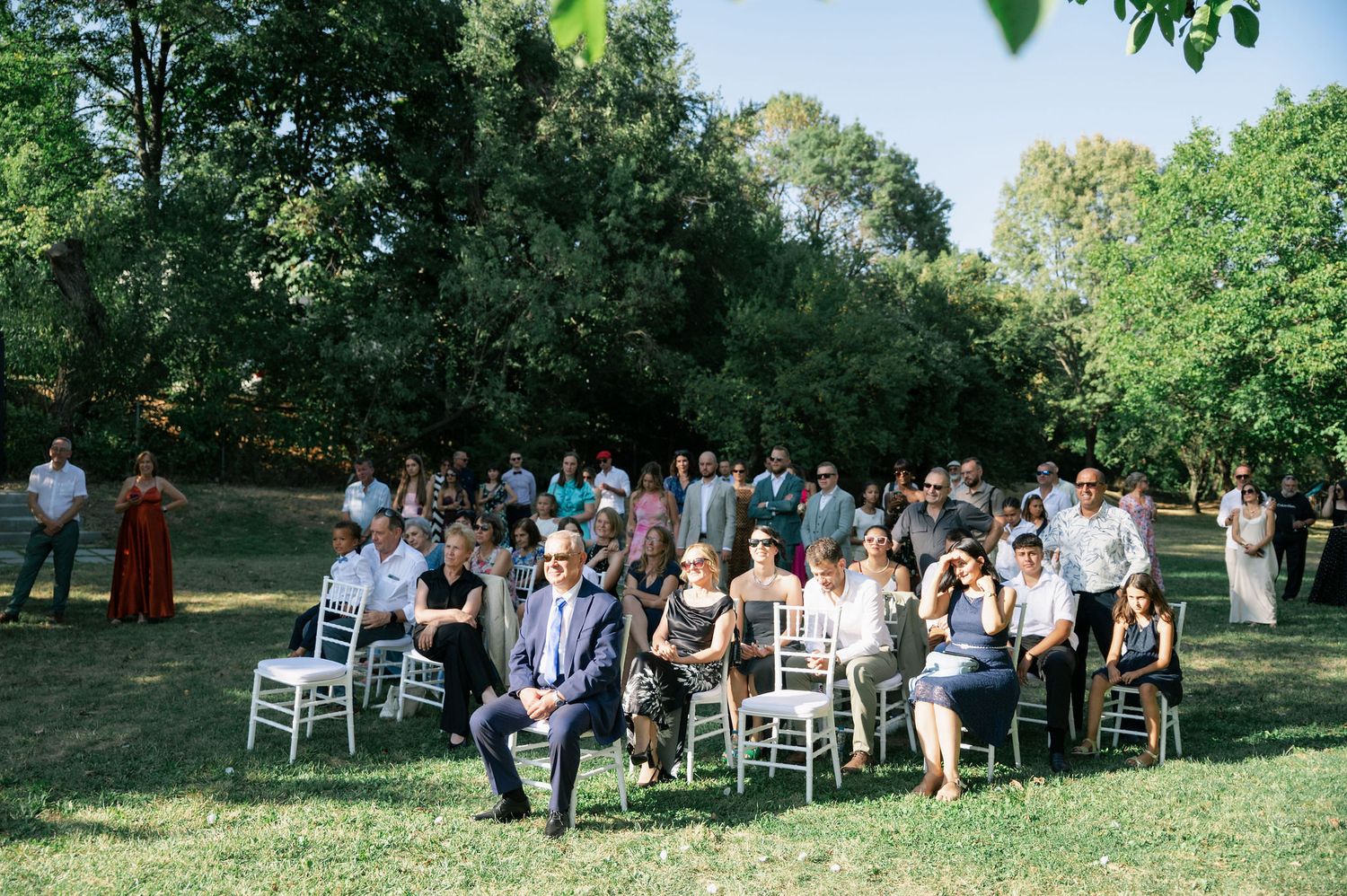 Wedding guests seated in white chairs on a grassy lawn during an outdoor ceremony.