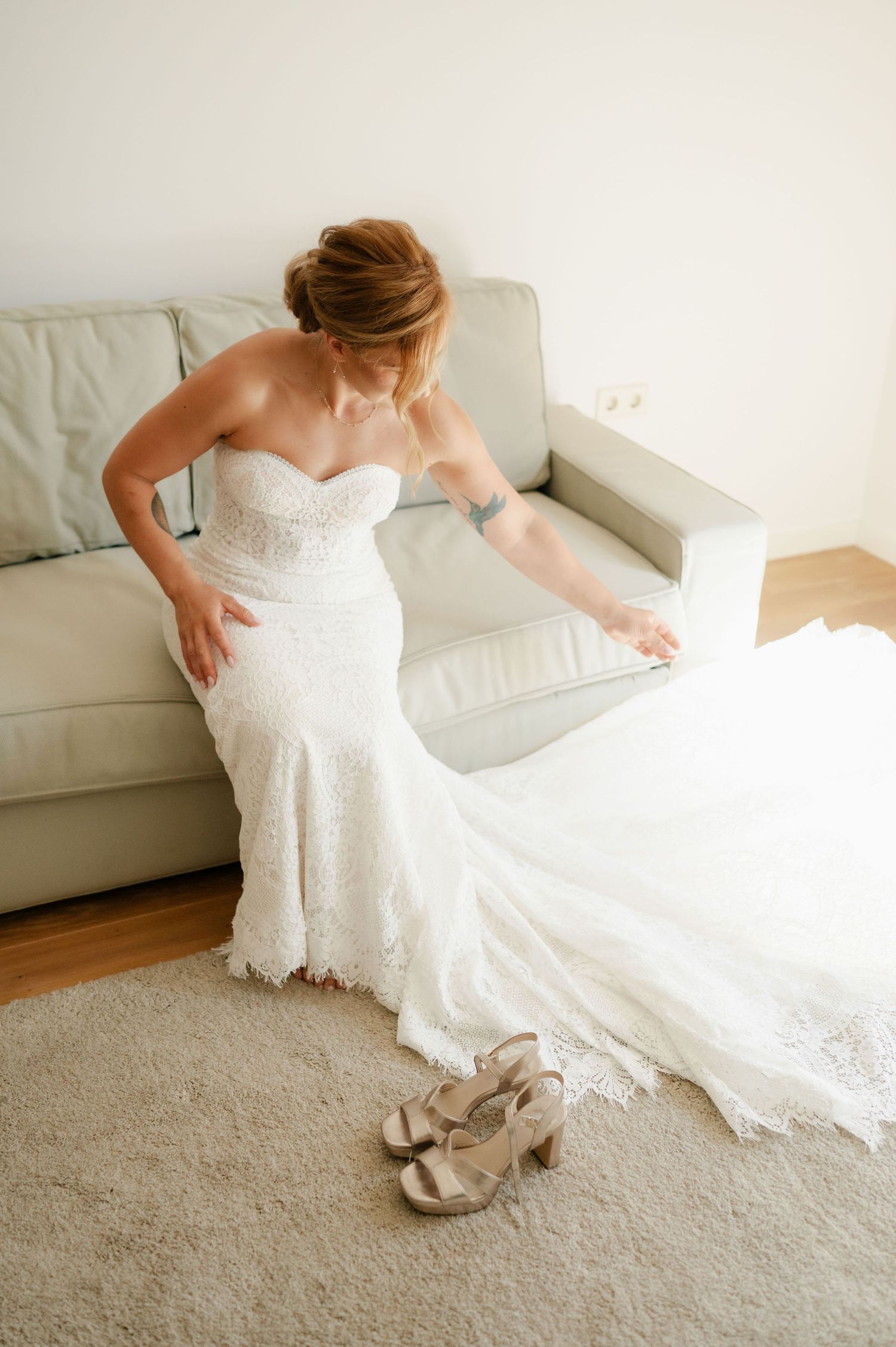A bride in a white wedding dress reaches down to adjust her shoes on a beige carpet.