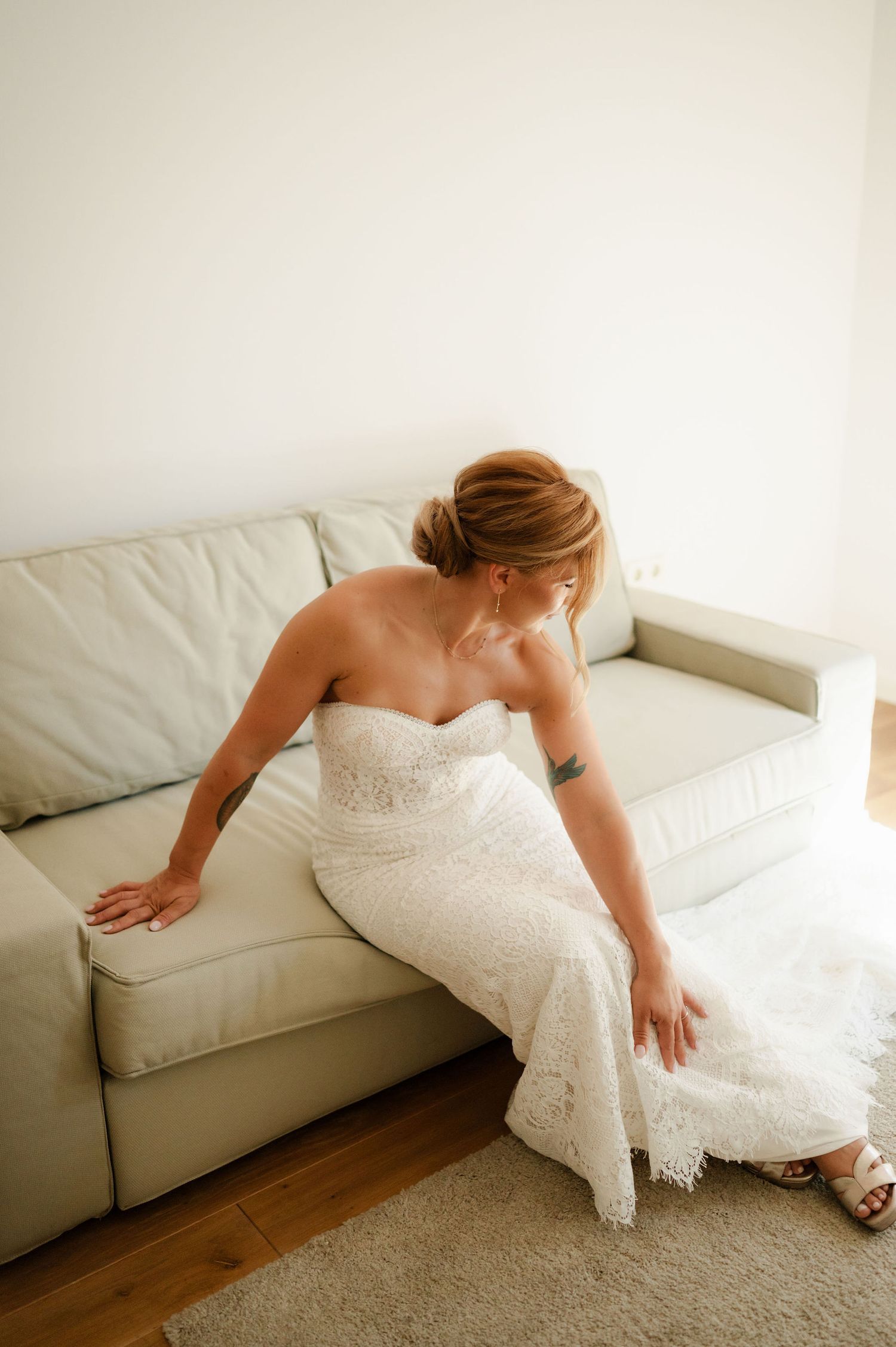 Someone in an elegant white bridal gown adjusts their position on a light colored sofa.