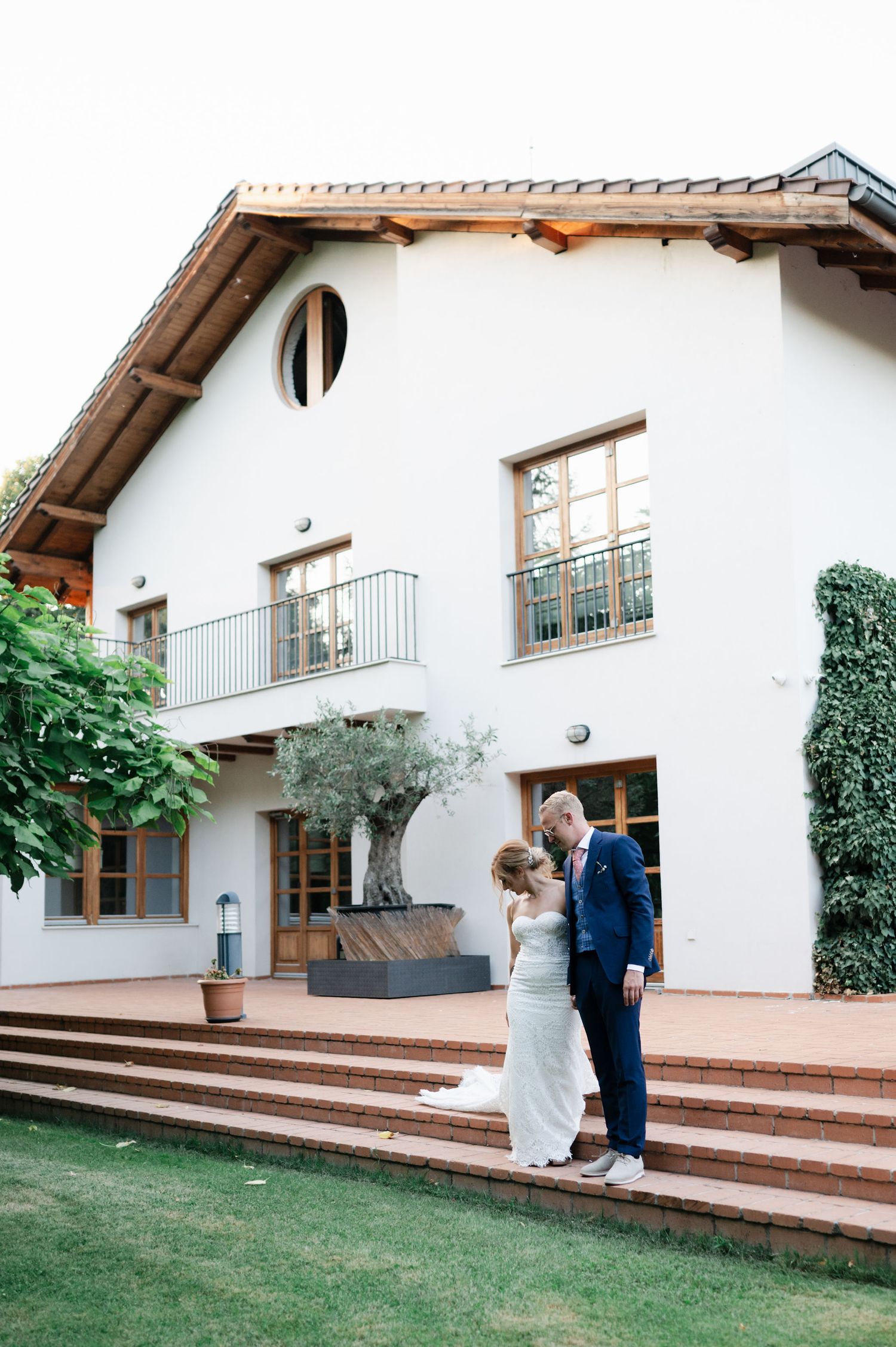 Newlyweds pose playfully on wooden steps outside a white Mediterranean-style villa.
