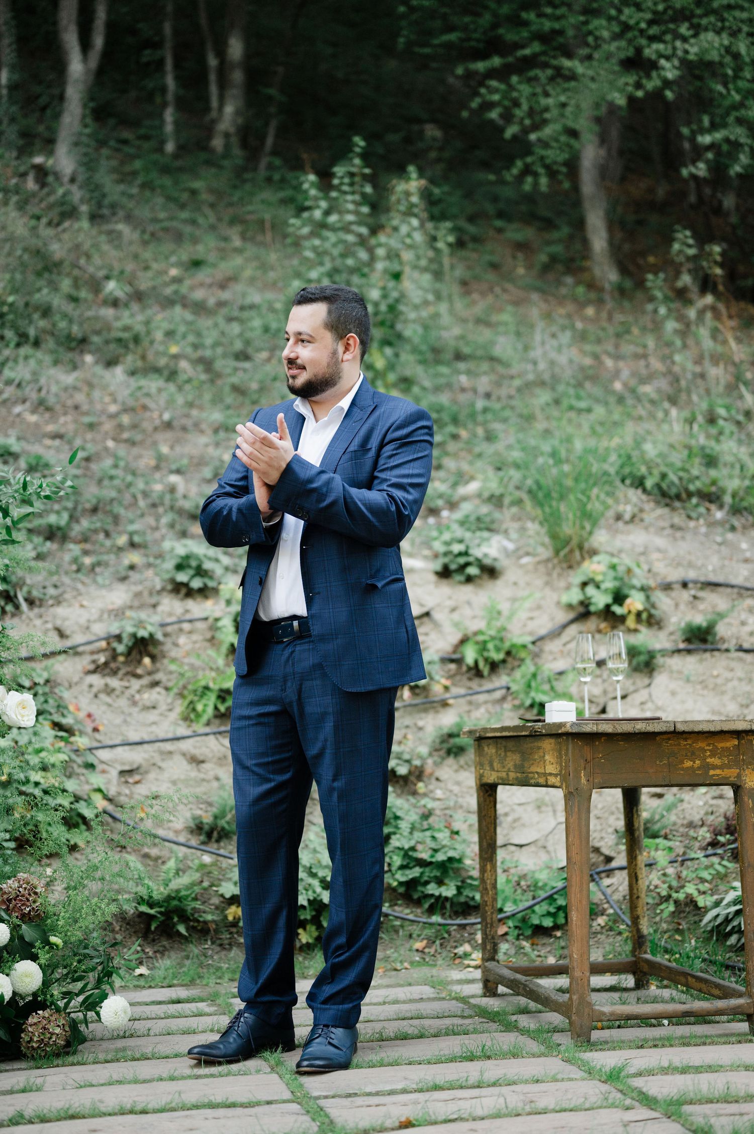 Person in navy suit stands on wooden deck during outdoor ceremony.