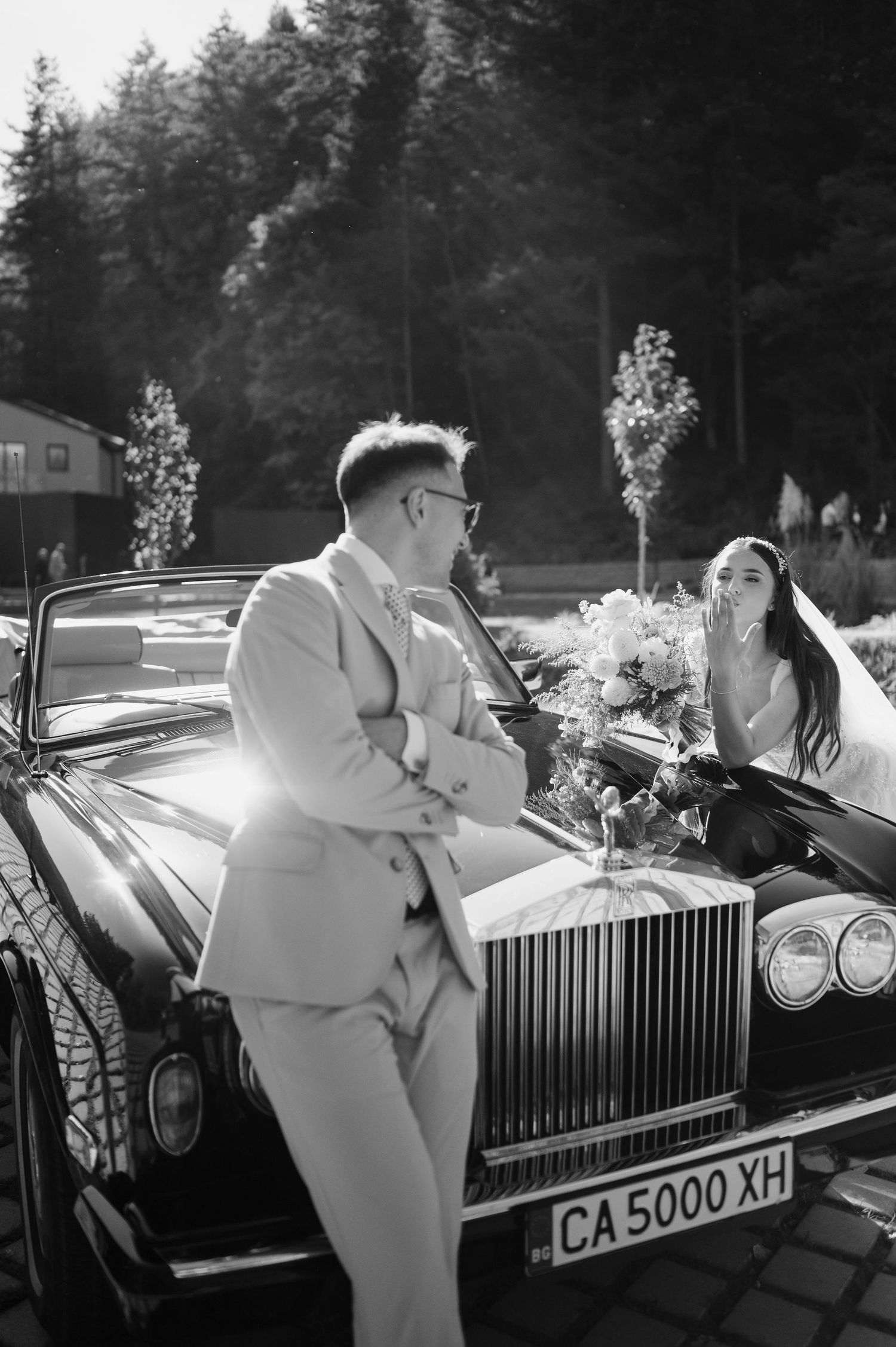 A couple shares a romantic moment beside a vintage Rolls Royce on a paved outdoor area.