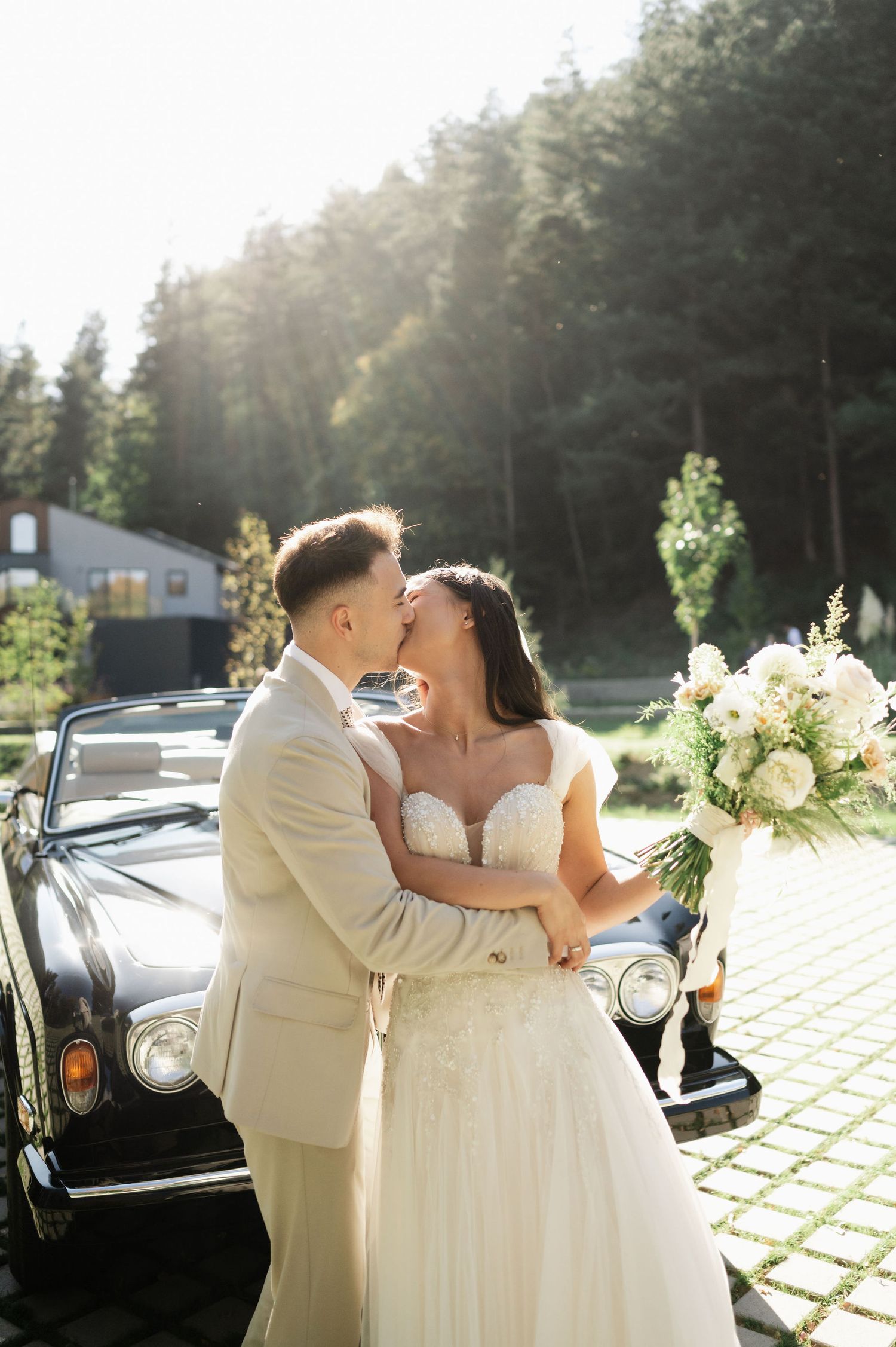 Newlyweds embrace in romantic moment with sunlit forest backdrop and classic car.