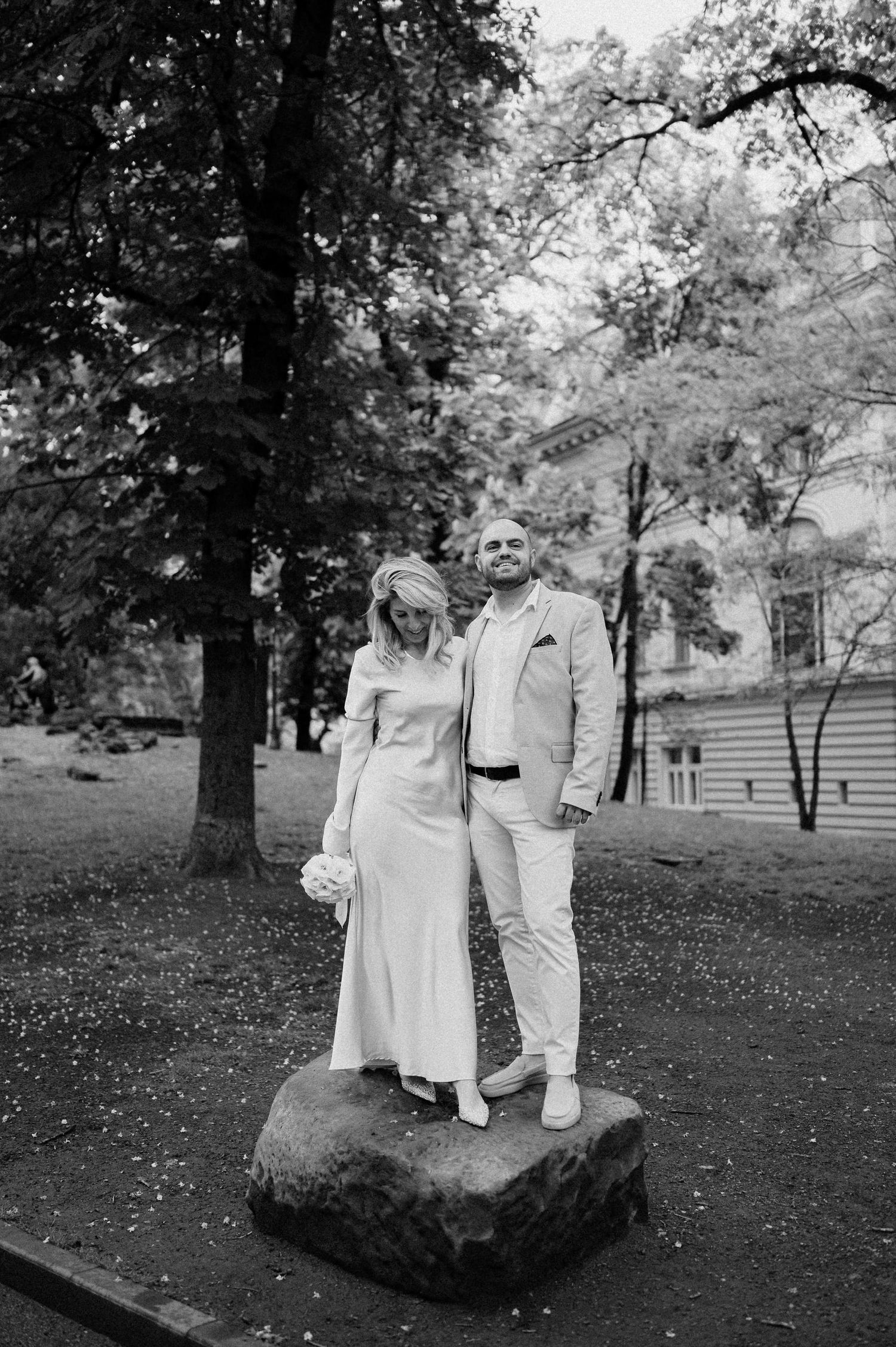 A couple in matching light outfits stands together on a large rock in a park setting.