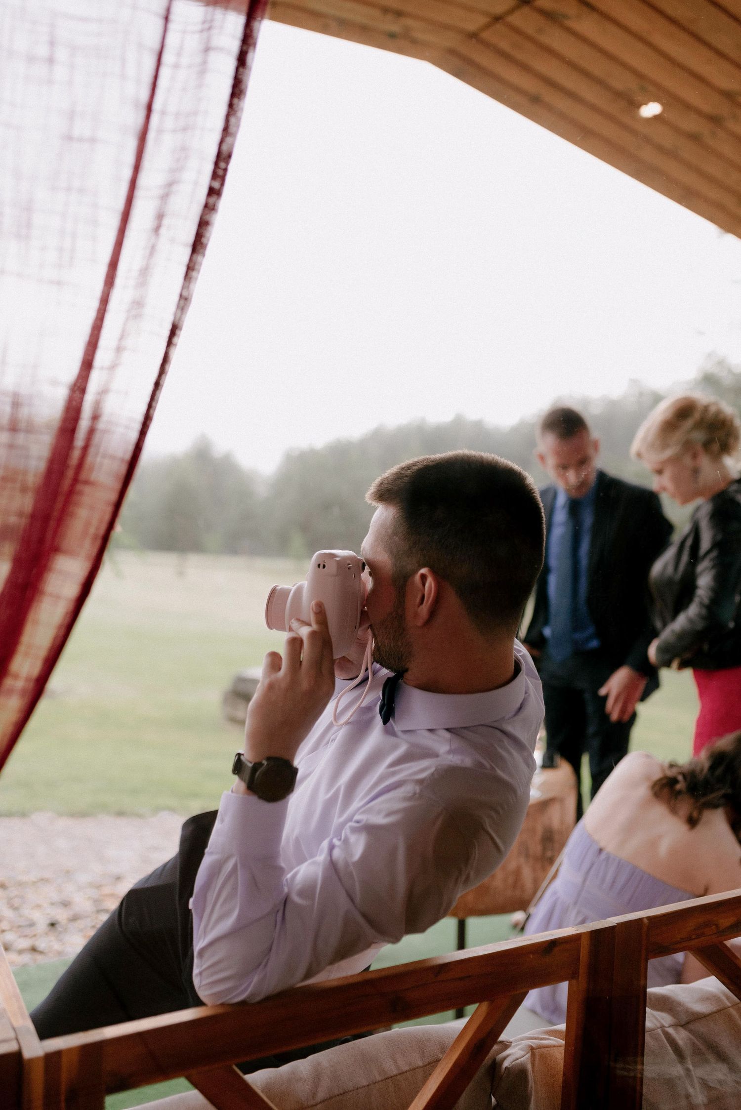 Guests seated on wooden benches watch an outdoor ceremony under a covered pavilion.