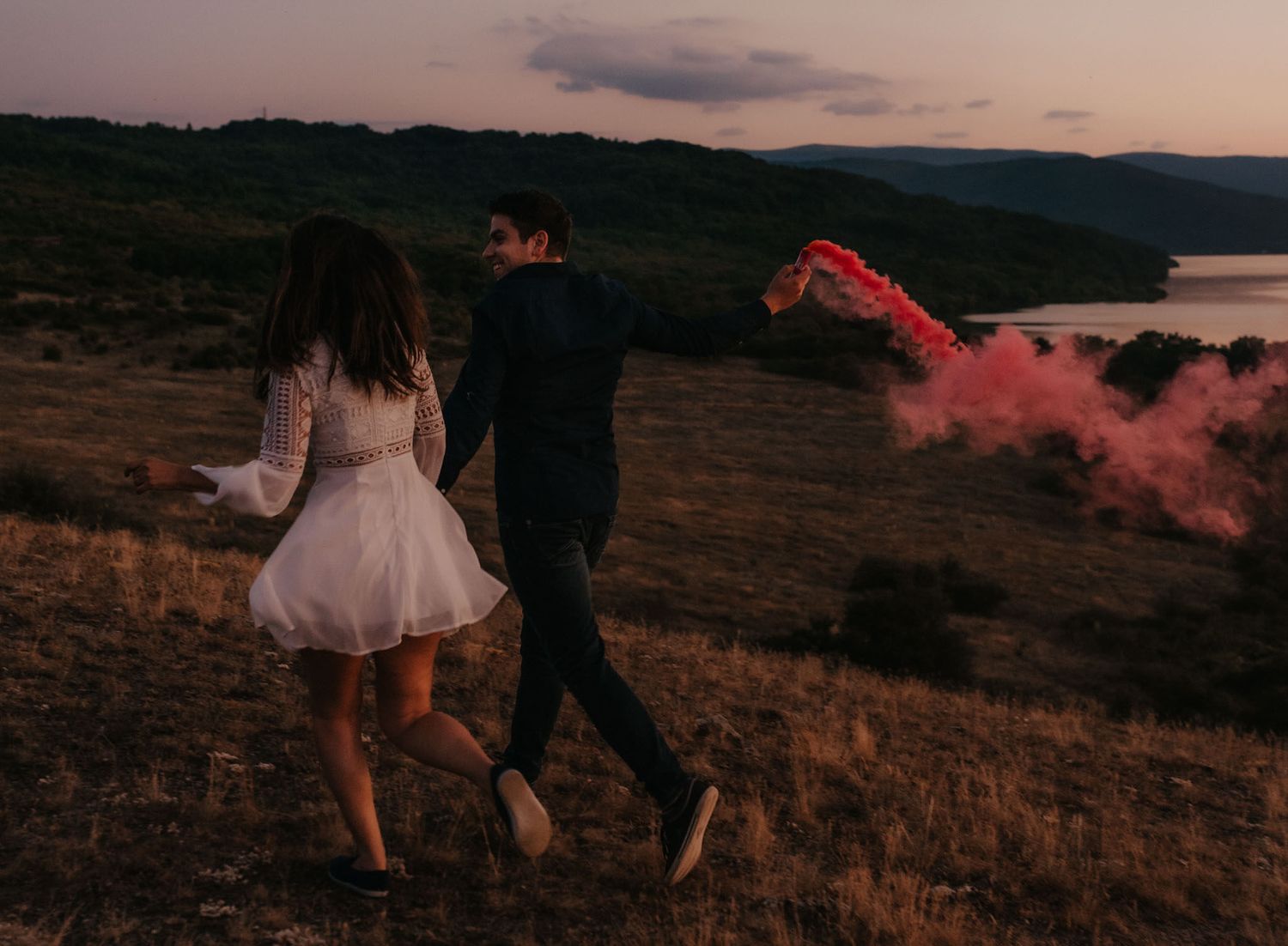 Two people walking along a hillside path at sunset with a lake vista in the background.