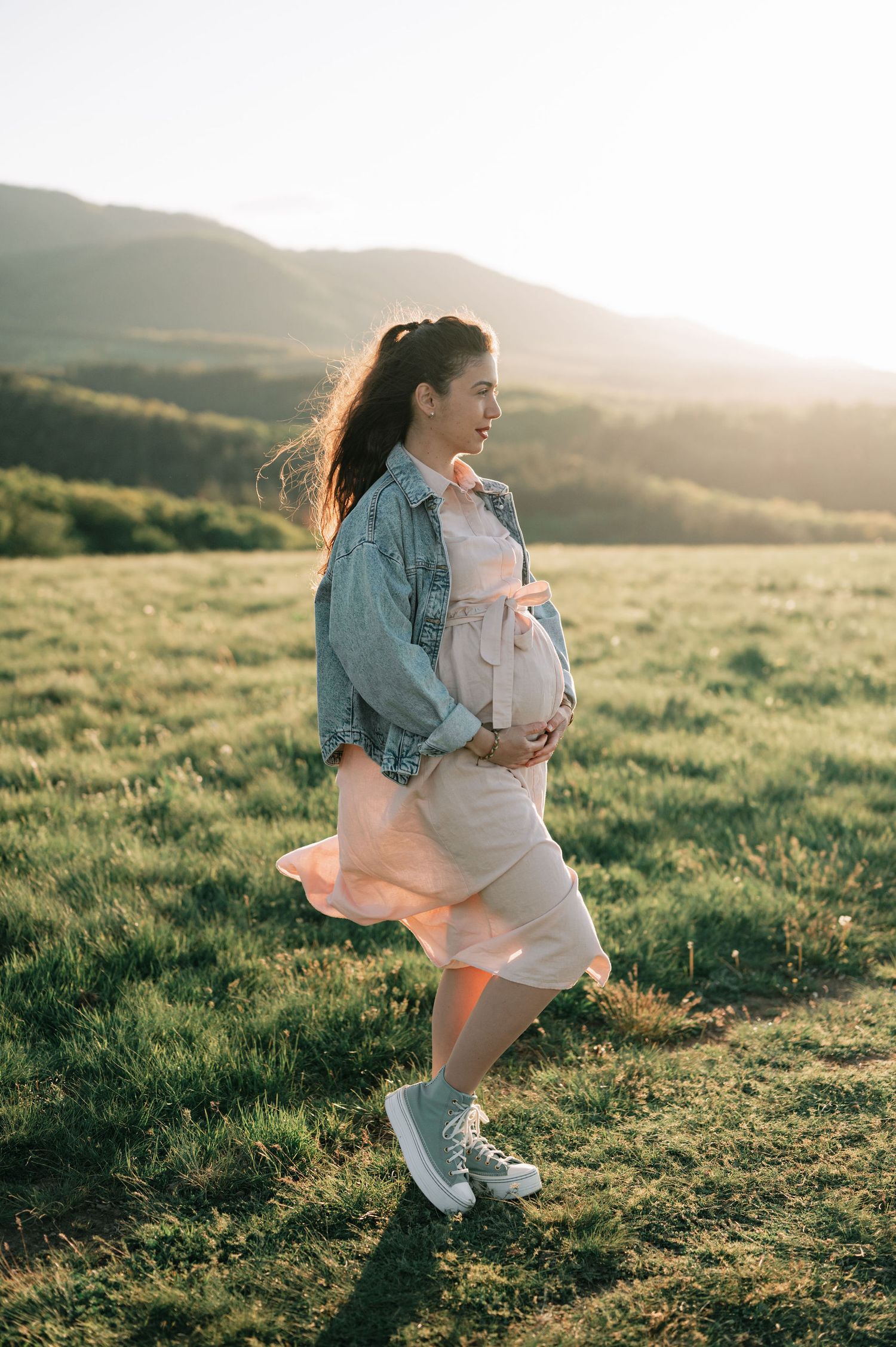 A figure dances joyfully in a flowing white dress and denim jacket across a sunlit hillside meadow at golden hour.