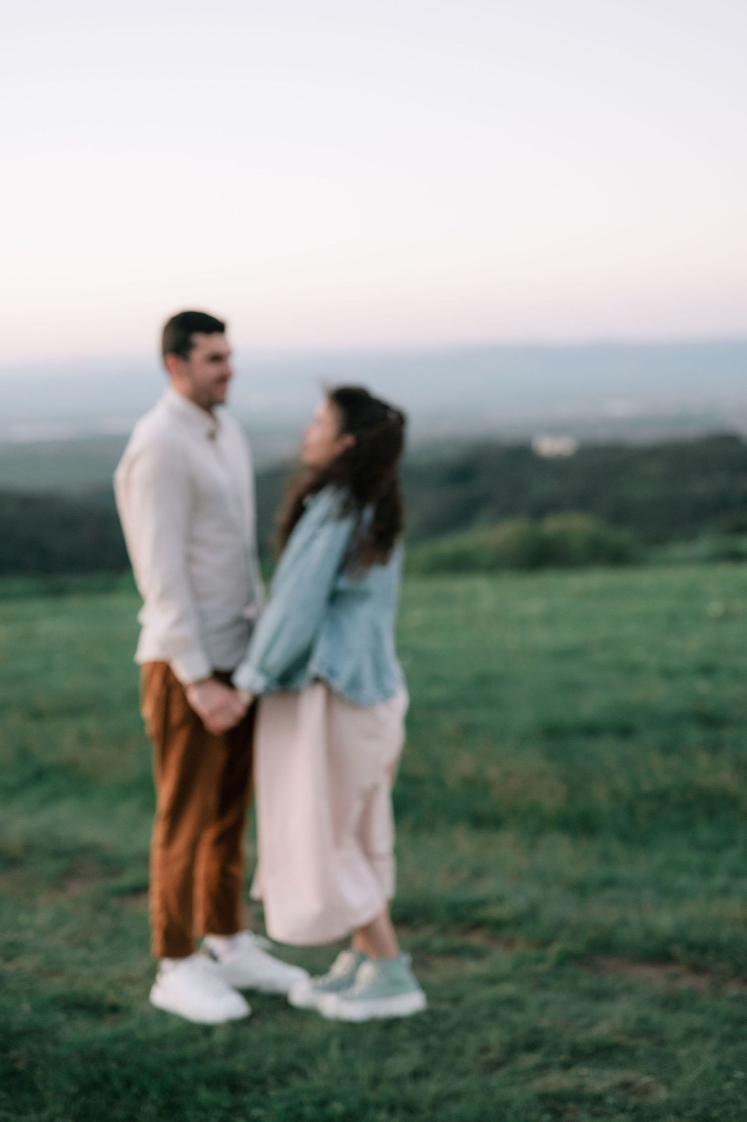 A couple in white and brown outfits share a tender moment together on a grassy hilltop at dusk.