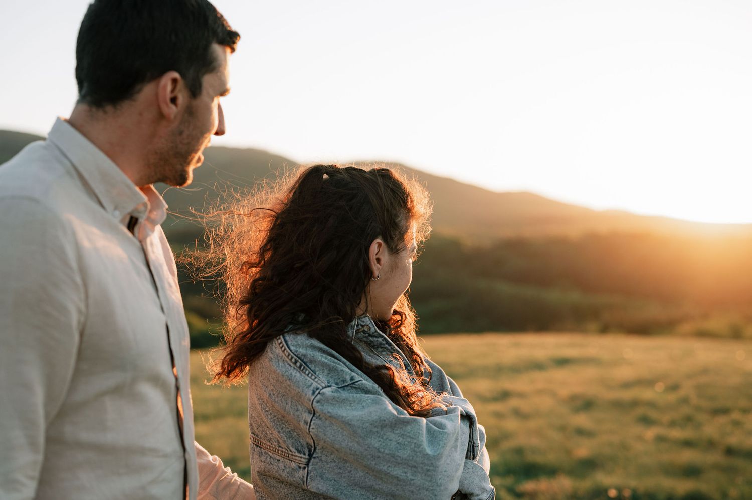 A couple stands together in a golden field at sunset with warm sunlight illuminating the peaceful countryside scene.