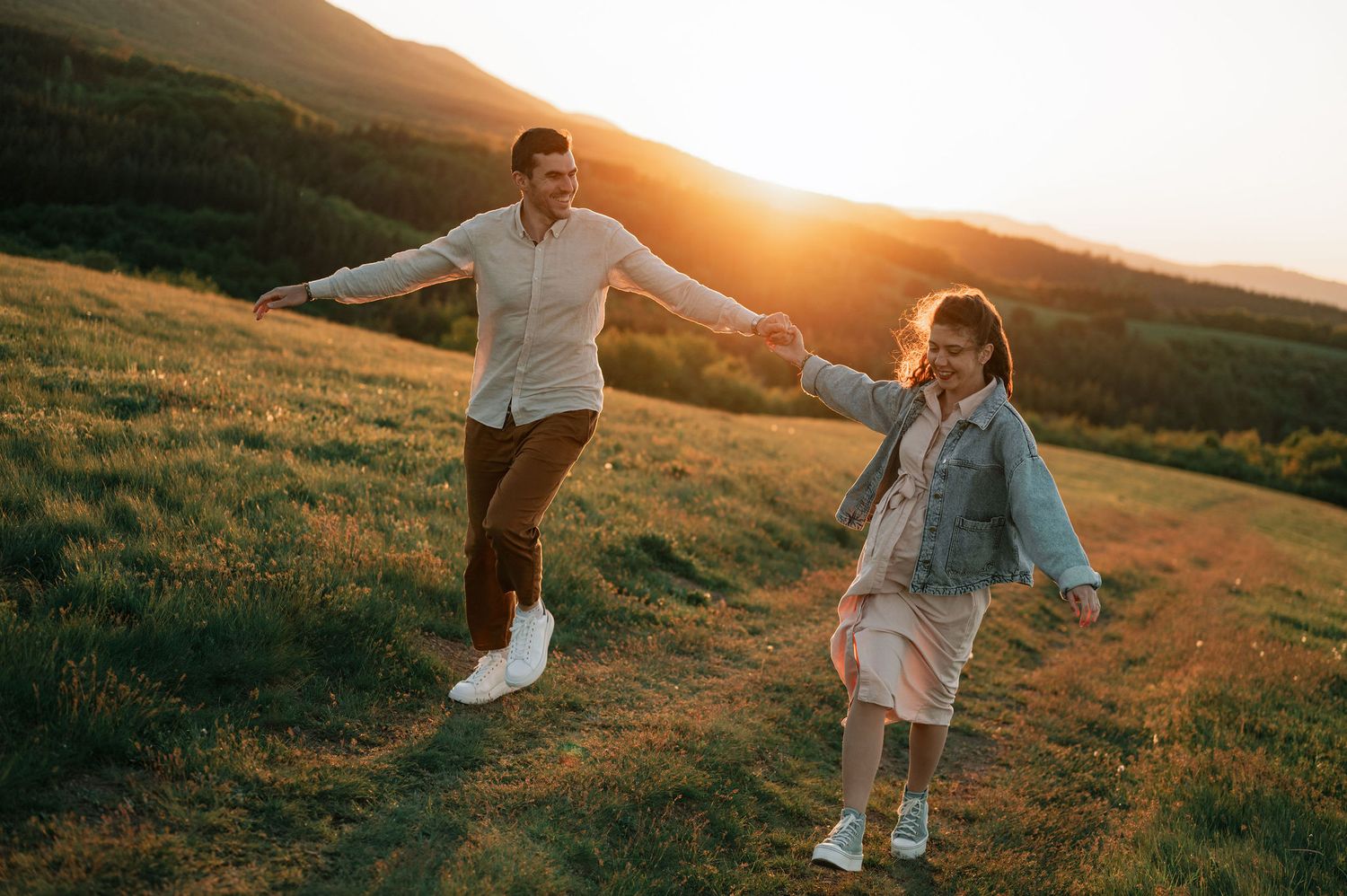 Two people playfully run and twirl on a hilltop meadow at sunset.