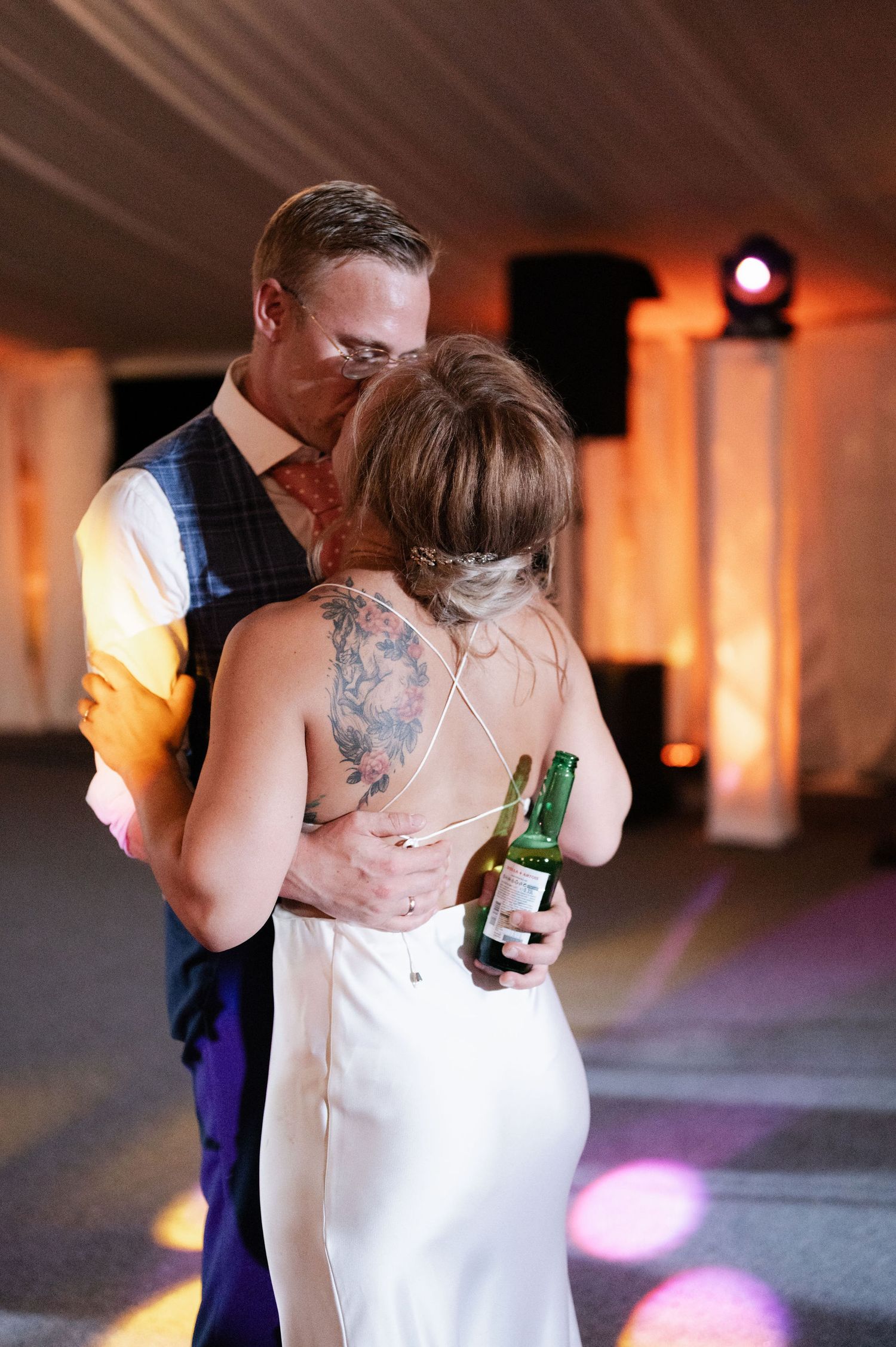A couple slow dances together at a dimly lit event venue while one holds a bottle of beer.