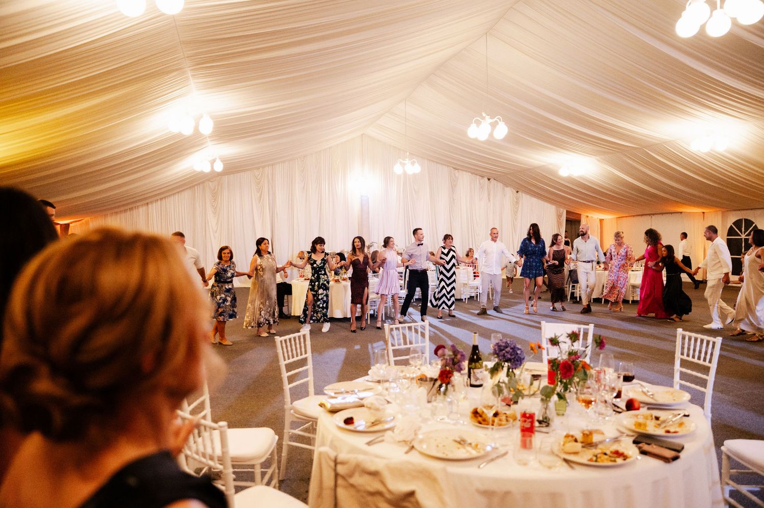 Wedding reception guests seated at elegantly decorated round tables in a white draped tent venue with warm lighting.