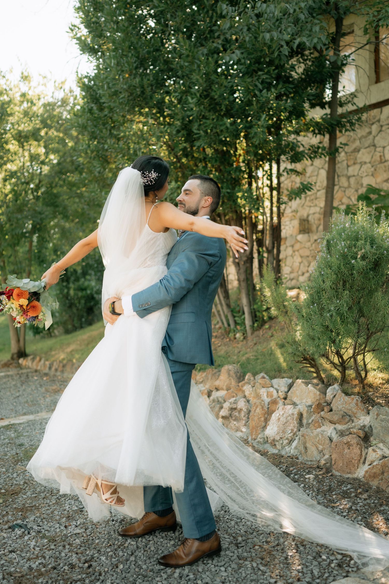 Romantic couple dances together outdoors in formal wedding attire with flowing veil and natural stone backdrop at sunset.