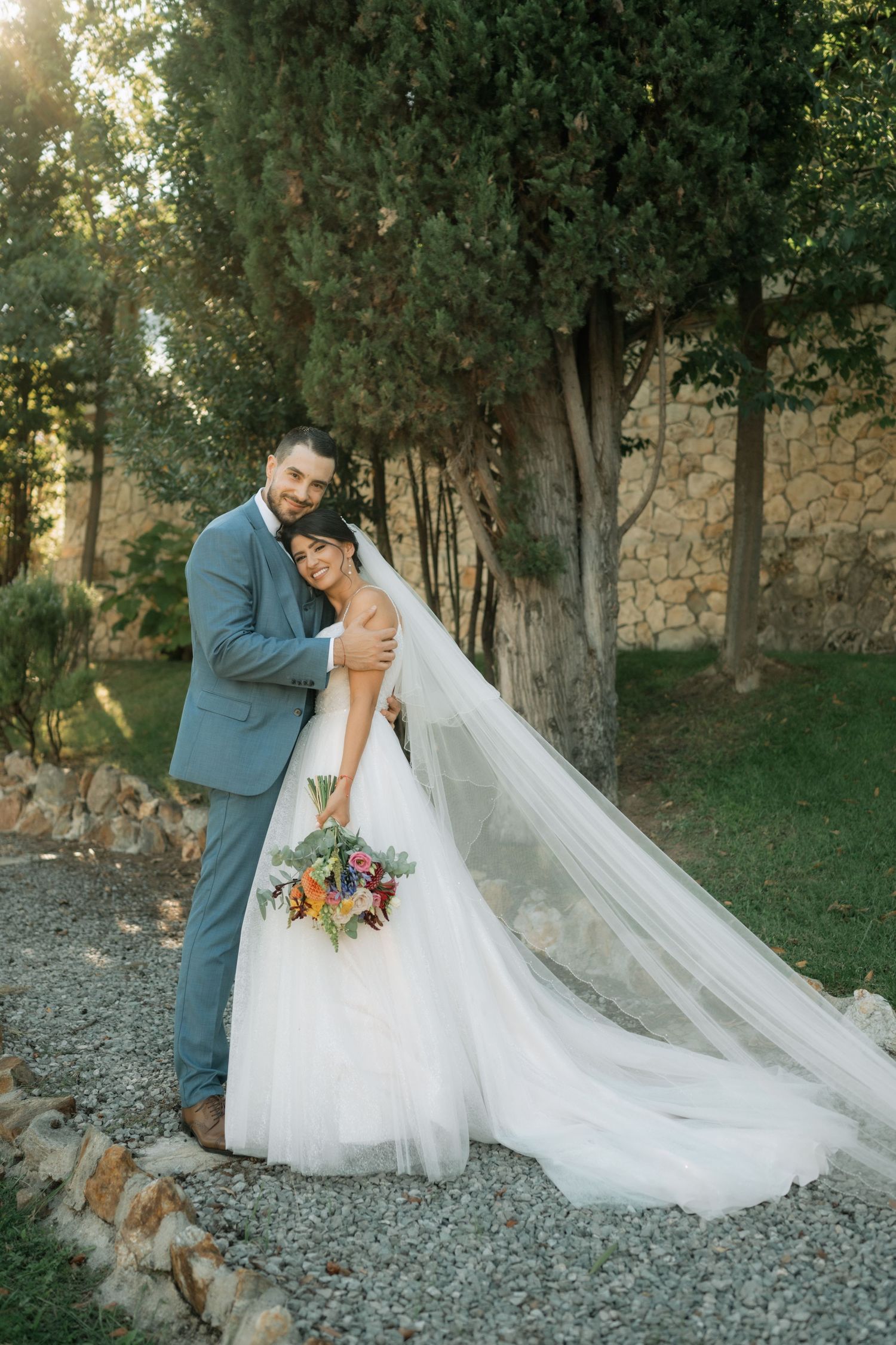 A romantic wedding couple shares an intimate moment on a stone path surrounded by lush trees and greenery.