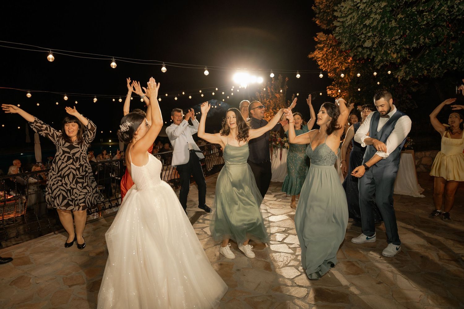 Wedding guests dance under twinkling string lights at an outdoor evening reception celebration.