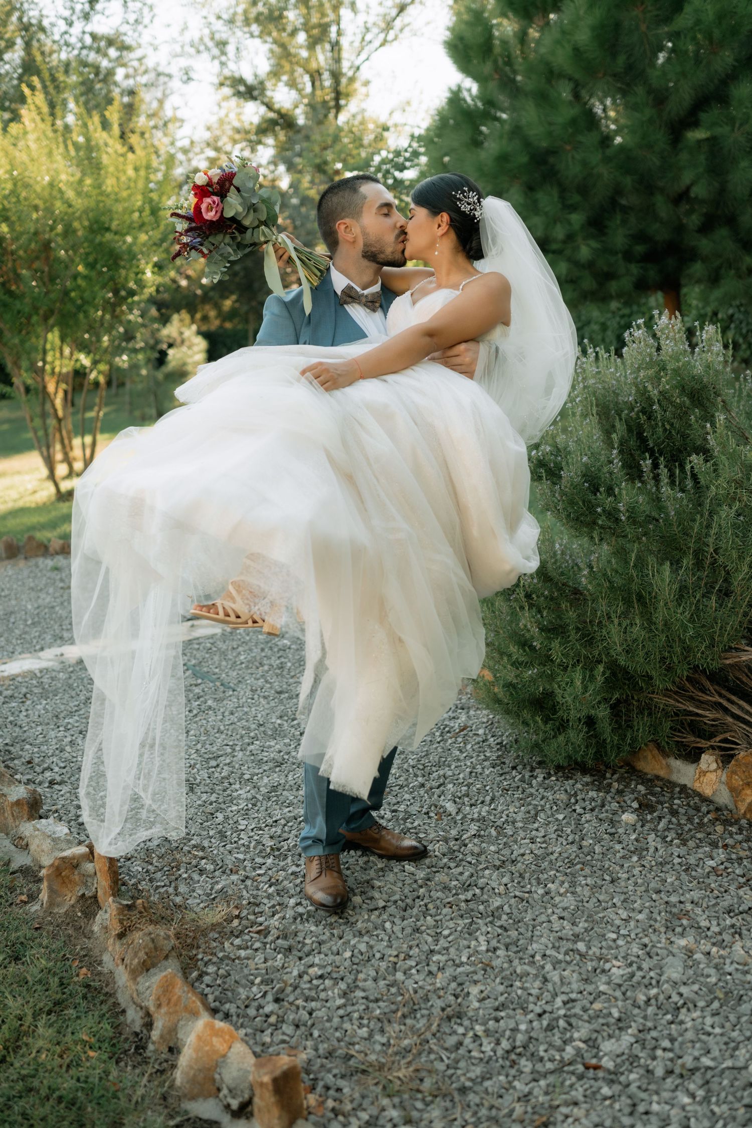 A romantic wedding moment captured on a gravel path with evergreen trees and natural landscaping in the background.