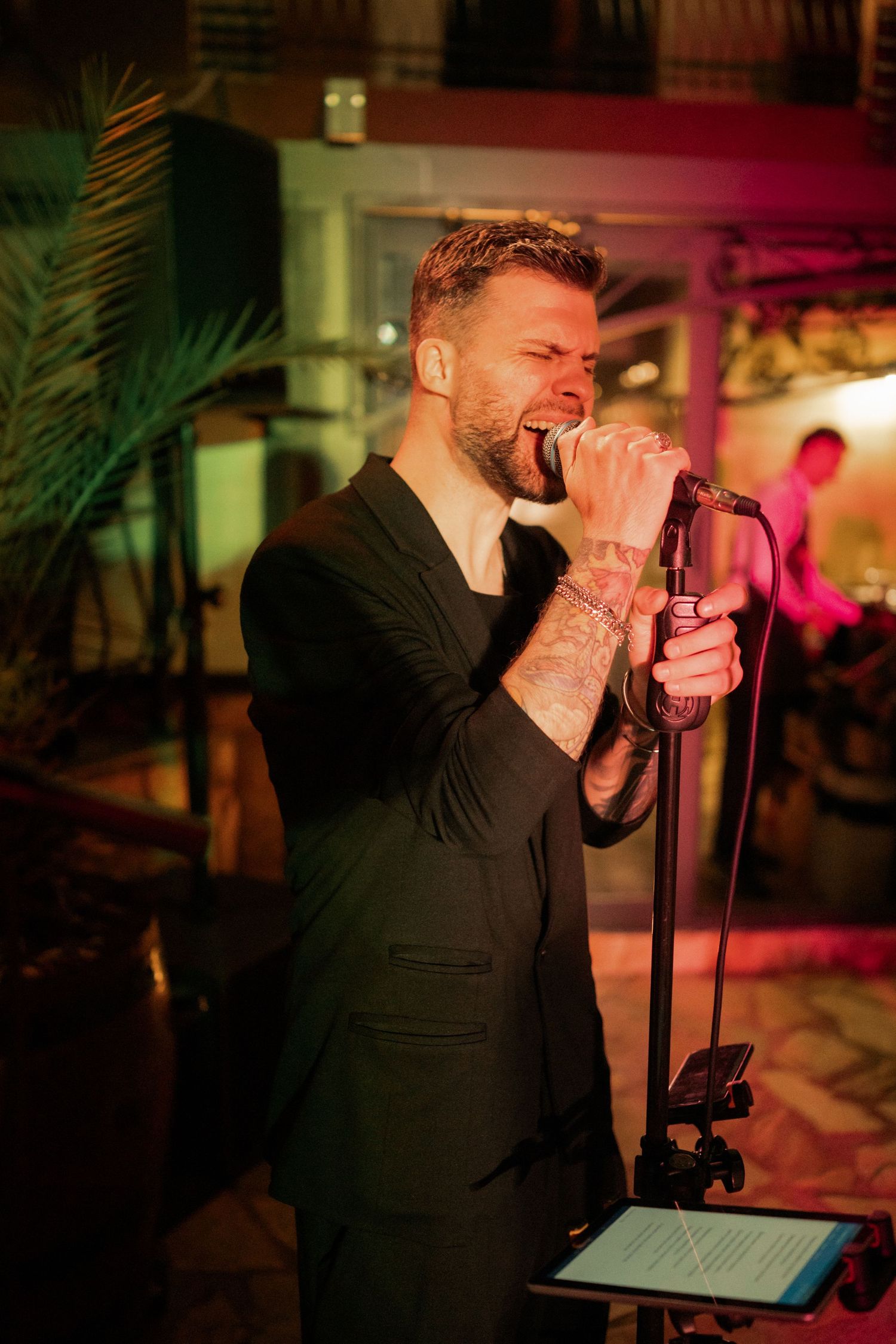 A singer in a black suit performs passionately on stage with a microphone under warm lighting at an indoor venue.
