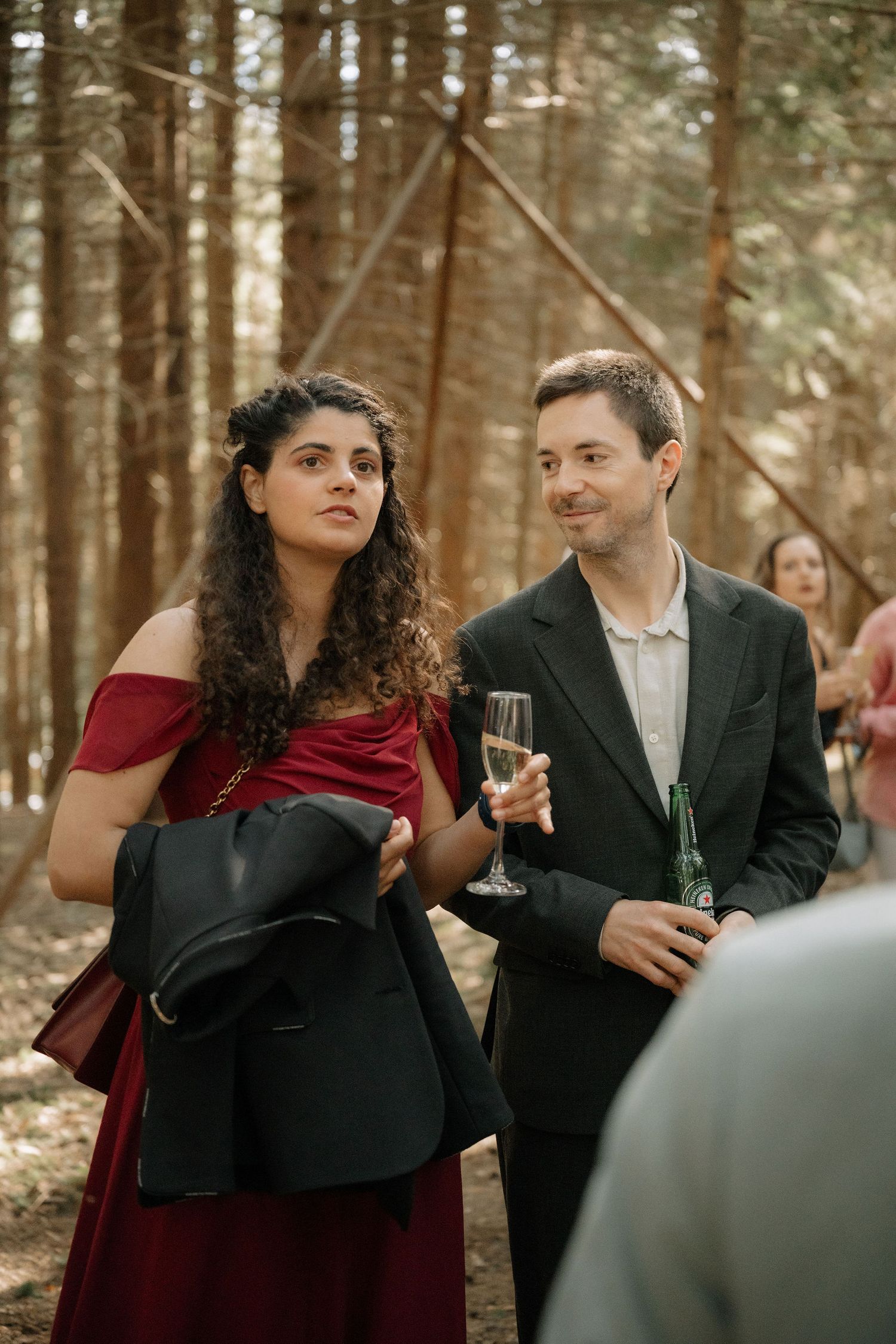 Couple in formal attire holding drinks at an outdoor woodland wedding reception among tall pine trees.