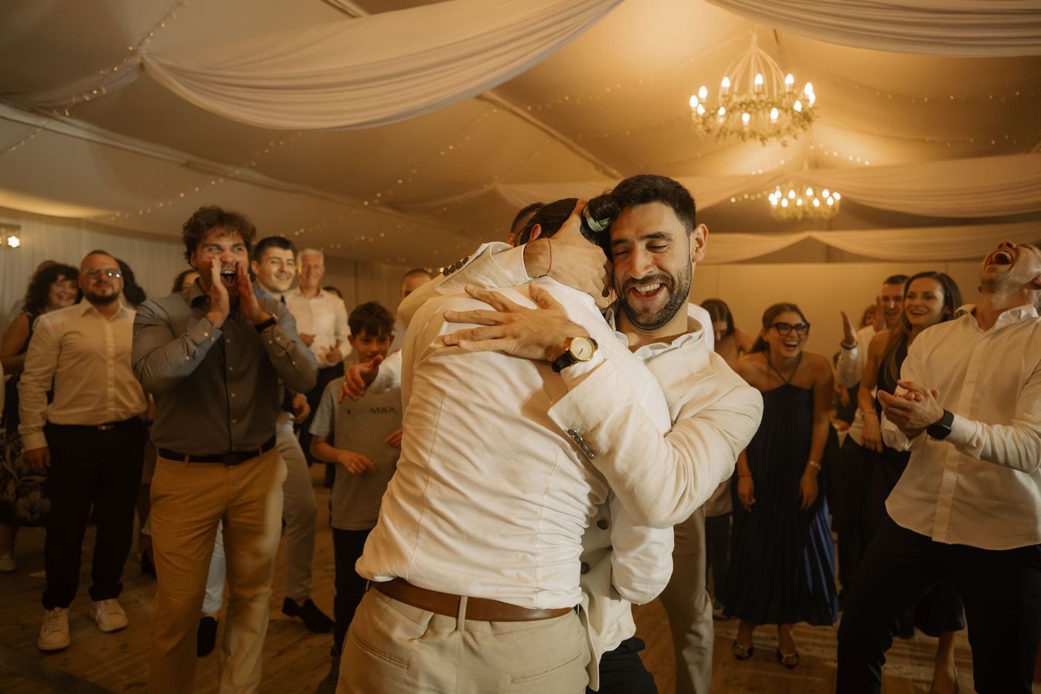 Two grooms share a joyful embrace on the dance floor surrounded by cheering wedding guests.