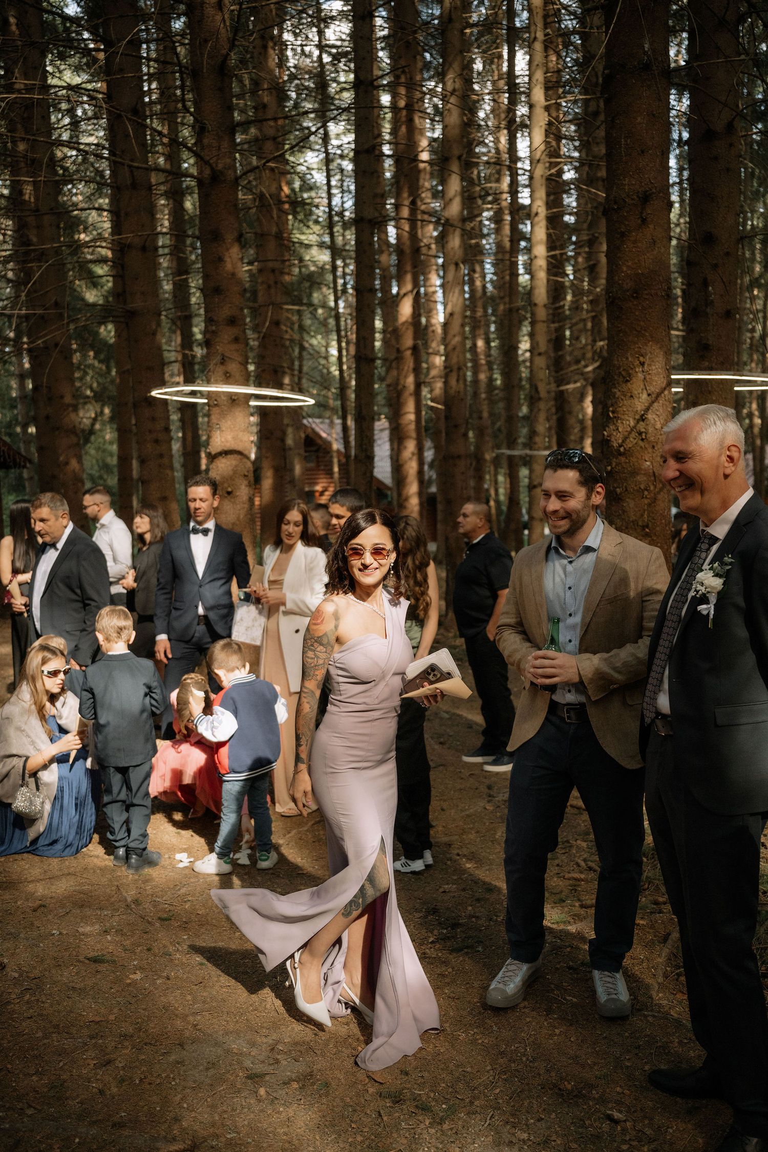 Elegant wedding guests socializing in a forest venue, woman in mauve gown with sunglasses walks confidently.