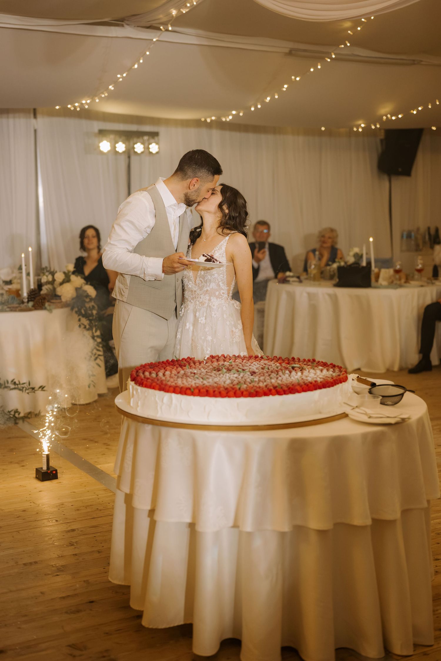 Bride and groom kissing while cutting their wedding cake decorated with red strawberries at a romantic reception venue.