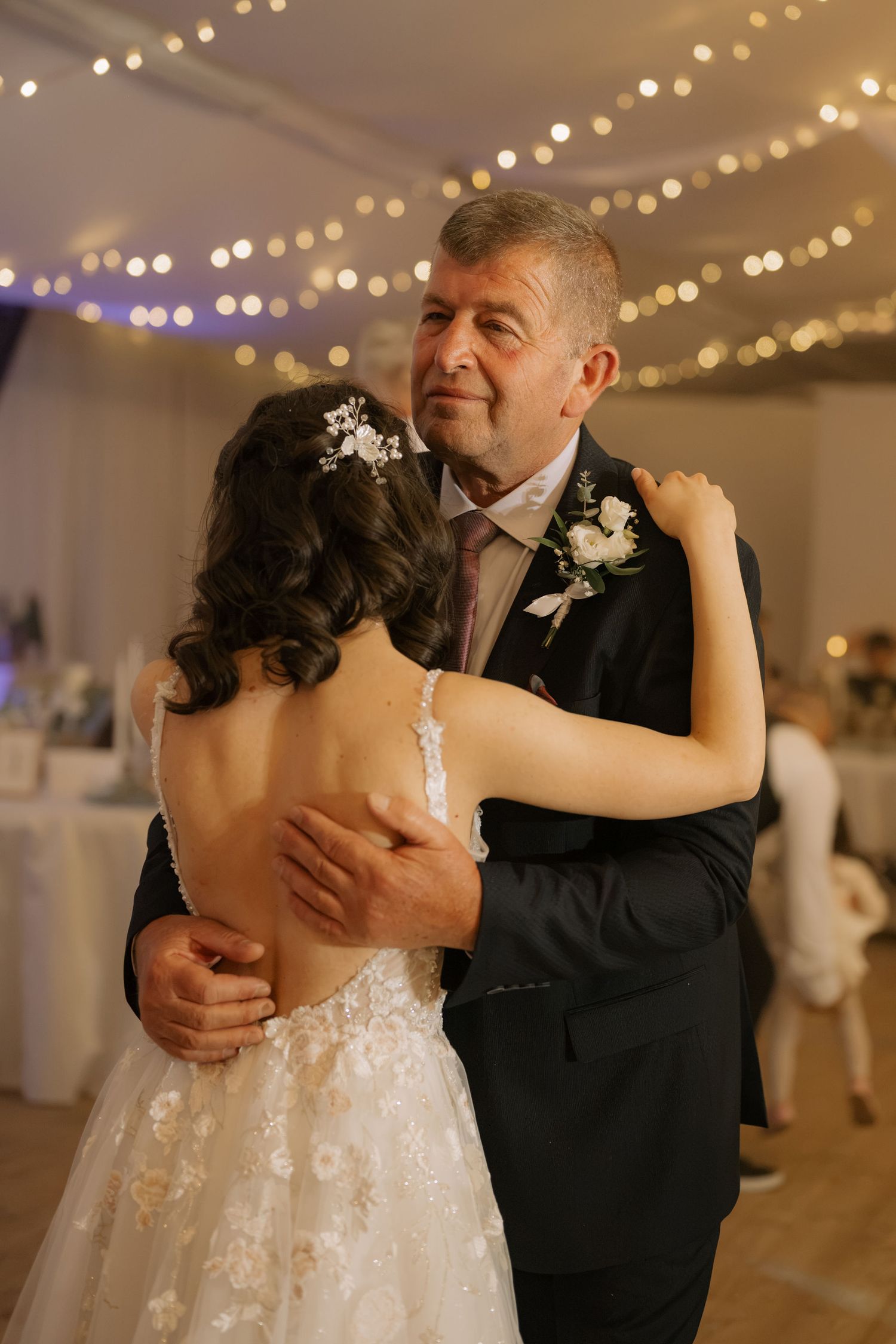 A bride and her father share an emotional first dance at a wedding reception with warm fairy lights glowing in the background.