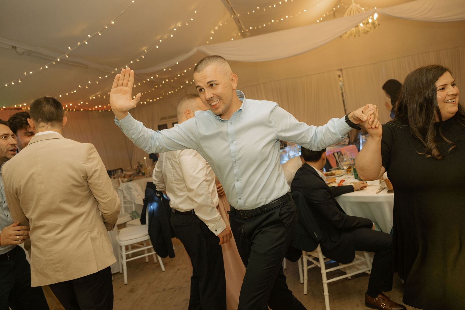 Man dancing joyfully at a wedding reception under string lights in a decorated tent venue.