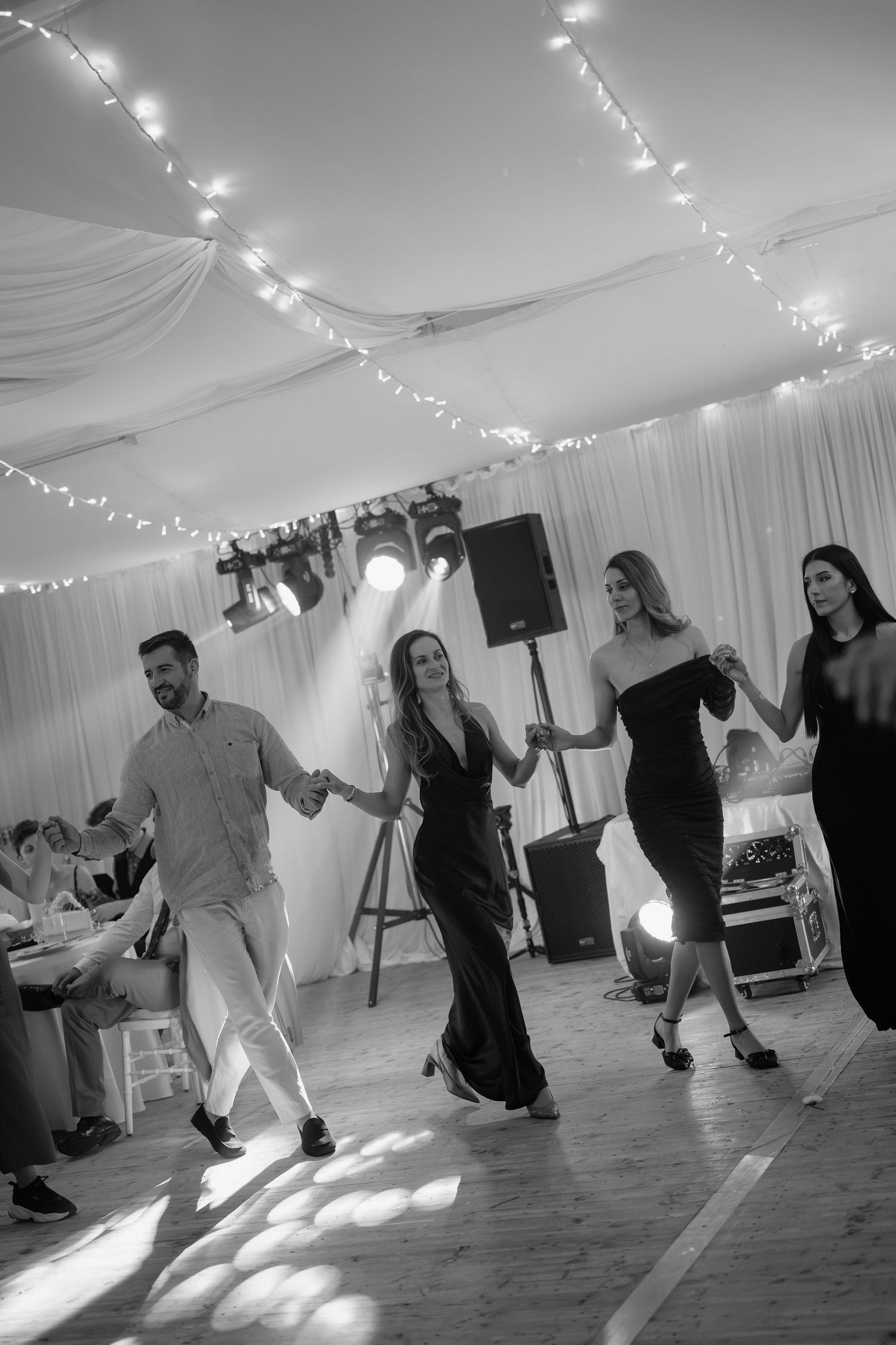 Black and white photo of four elegantly dressed guests dancing together under fairy lights at a wedding reception.
