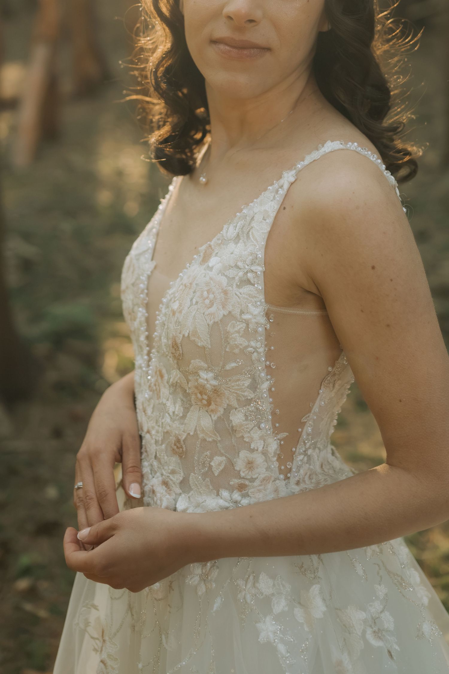 Bride in floral lace wedding gown with deep V-neckline poses outdoors in soft natural light among trees.