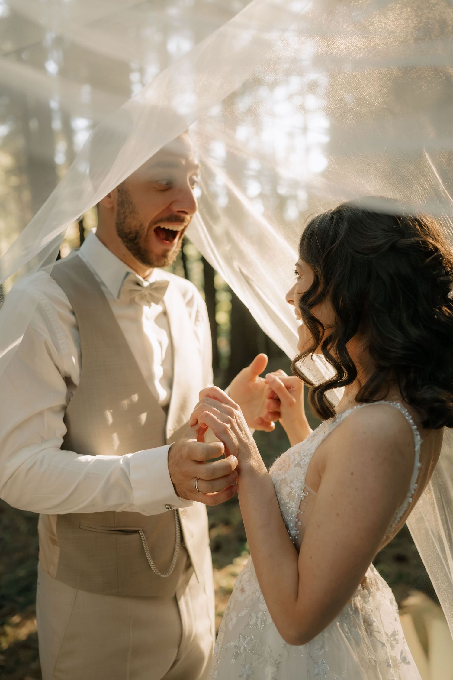 Joyful bride and groom sharing a laugh under a veil in a sunlit forest during their wedding ceremony.
