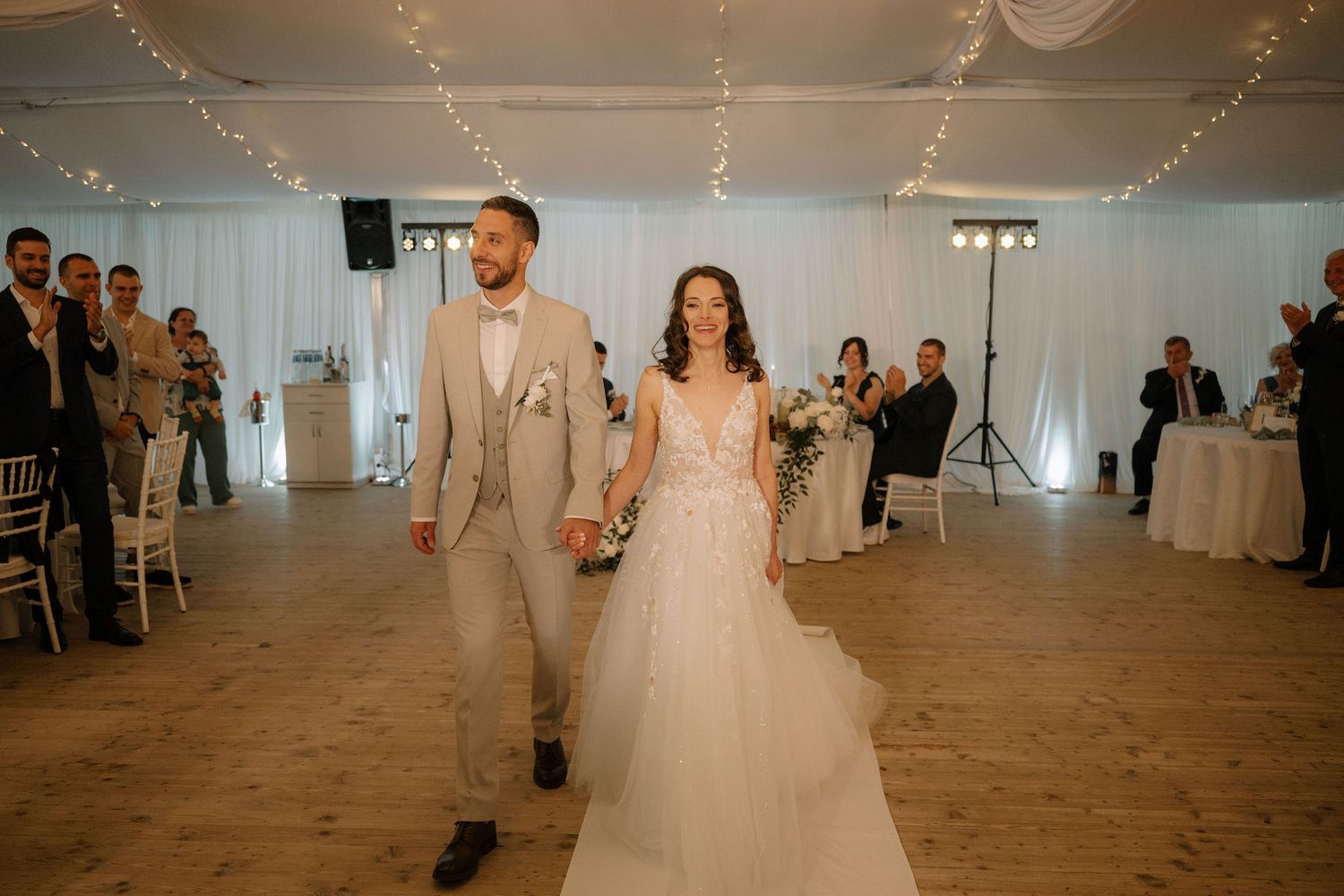 Bride and groom walk hand in hand at elegant indoor wedding reception with fairy lights and white draping.