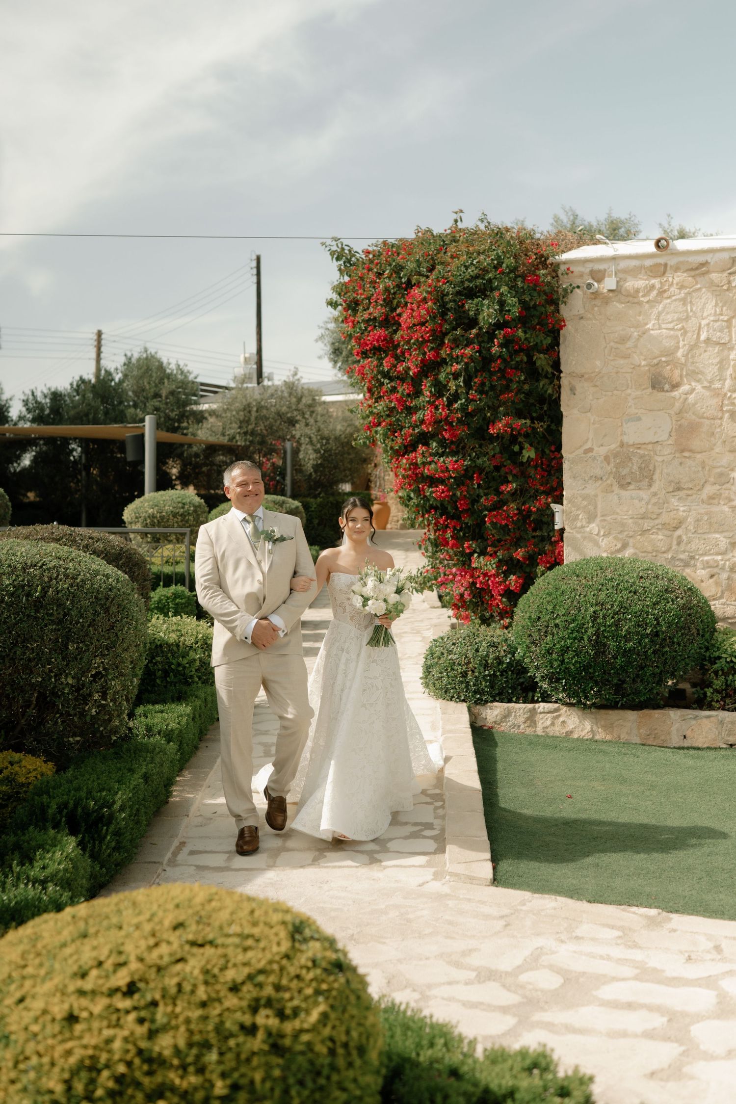 Bride and groom walk through a lush garden path with red climbing flowers and manicured hedges at an outdoor wedding venue.