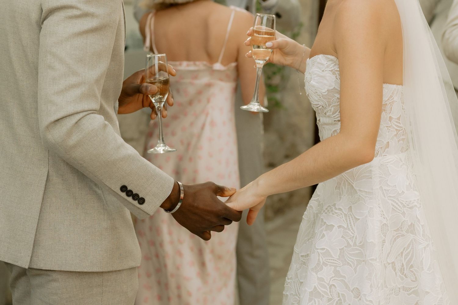 Interracial couple holding hands at wedding, bride in lace gown, guests toasting champagne in background.