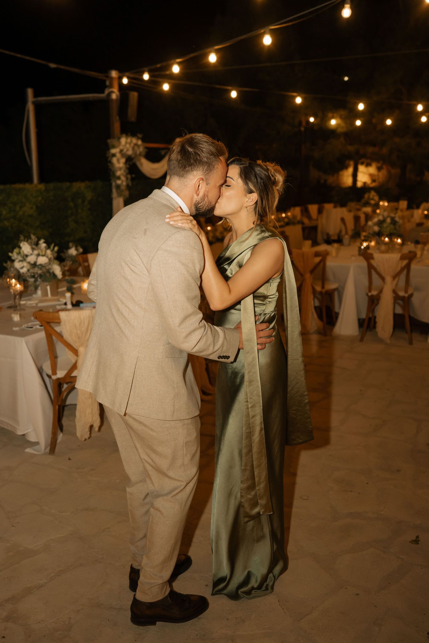Couple sharing a romantic kiss at an outdoor wedding reception with warm string lights glowing in the evening background.