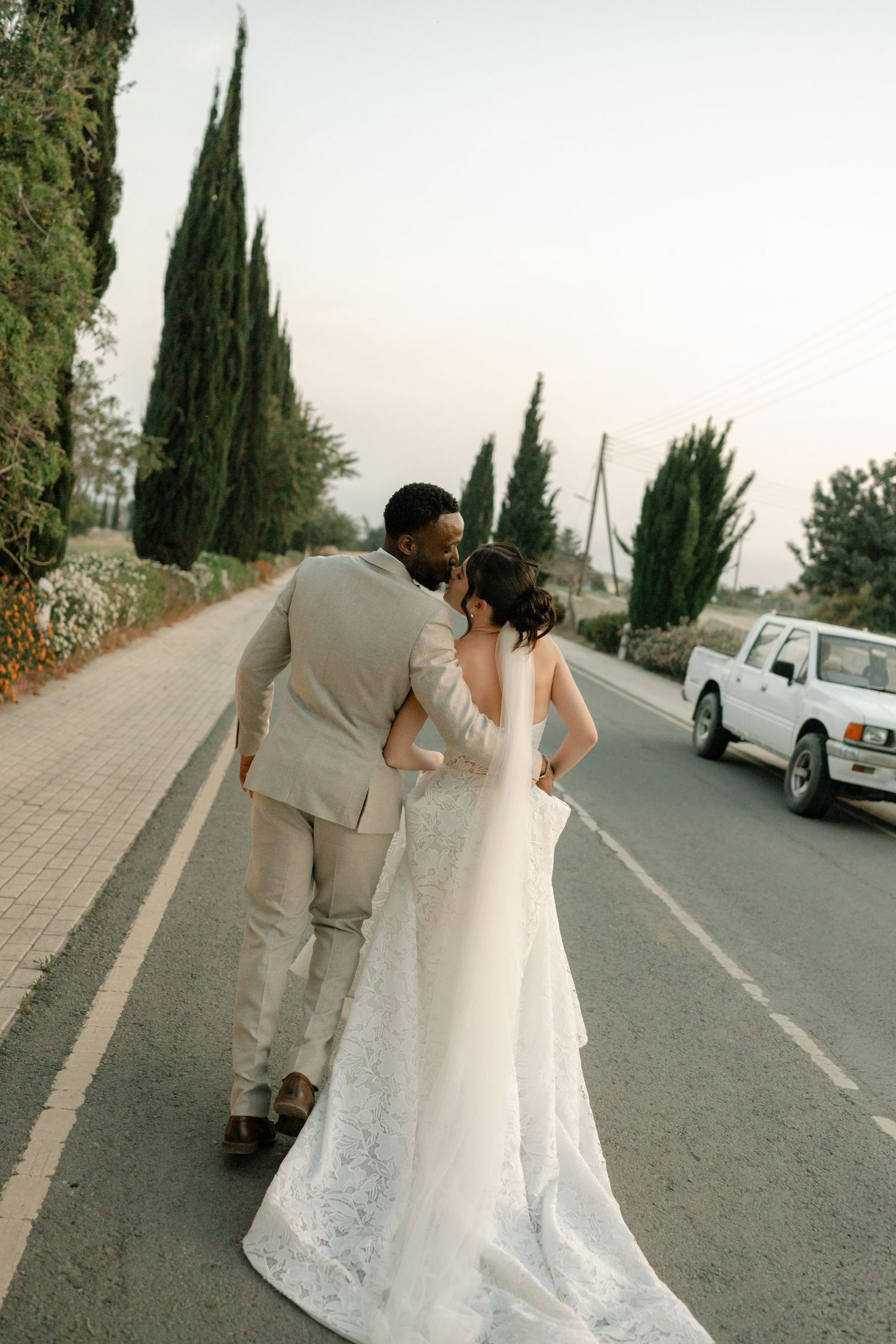 Bride and groom share a kiss on a scenic road lined with cypress trees, bride in lace gown, groom in beige suit.