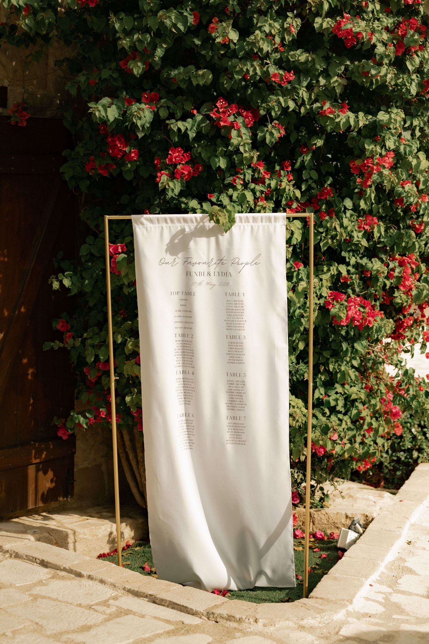 Gold frame wedding seating chart on white fabric displayed outdoors against lush green foliage with red bougainvillea flowers.